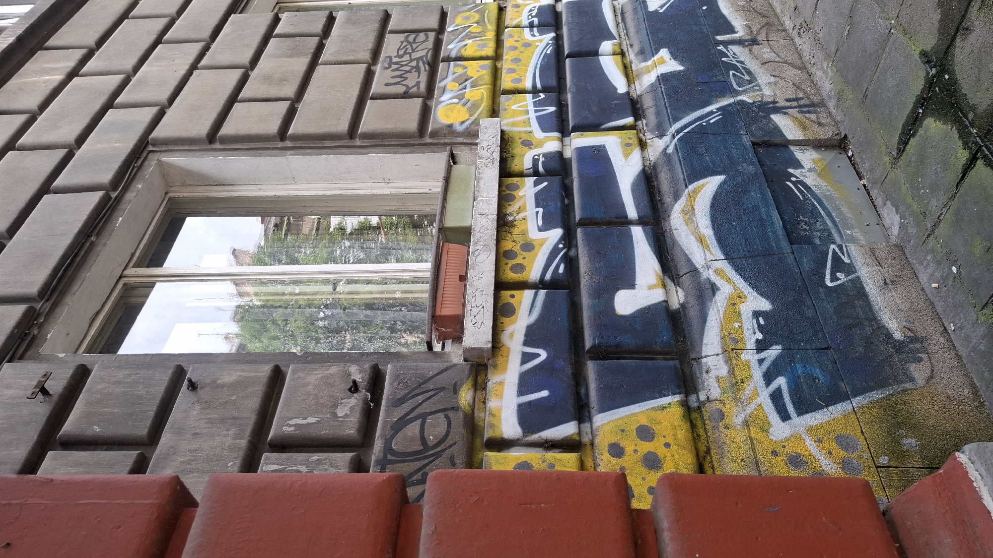 The image displays a close-up, low-angle view of a building facade with distinct textures and graffiti. The upper portion of the building features rows of rectangular, grey concrete tiles, possibly imitating brickwork, running diagonally from the top left. A window with a white frame is visible within this section, reflecting greenery and sky. Adjacent to the concrete tiles, on the right side, is a wall composed of vertical, rectangular blocks. These blocks are painted with bold graffiti featuring yellow, blue, and white spray paint. The yellow sections are marked with black dots, resembling a distressed or textured effect. White abstract lines and shapes are layered over the yellow and blue, creating a dynamic visual.
Below the window and concrete tiles, there are horizontal, rectangular wooden-like elements with a grey, distressed finish. Some of these elements have visible nails or hooks. At the very bottom of the image, a row of red, textured rectangular shapes, possibly painted concrete steps or a railing, frames the scene.
The graffiti suggests an urban environment, and the combination of aged building materials with vibrant street art creates a contrast. The overall impression is one of an exterior wall in a city, likely in a public or accessible area. There is no clear indication of the time of day or weather, though the reflections in the window suggest daylight. No discernible text is visible in the image. The context provided states the location is Sofia, Bulgaria.