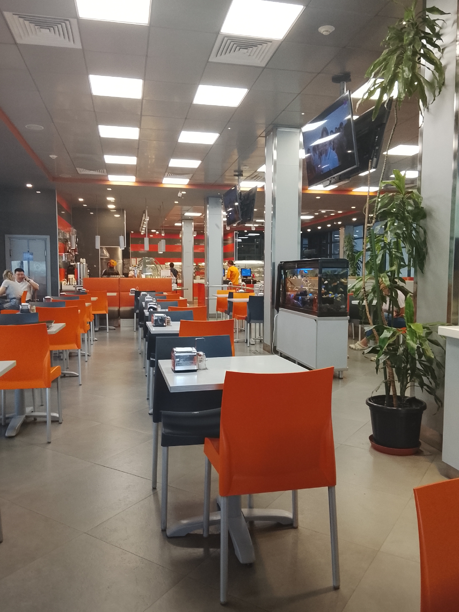 The image depicts the interior of a modern cafeteria or food court, likely located in Almaty, Kazakhstan. The environment is well-lit, with numerous ceiling lights casting a bright glow on the tiled floor and suspended ceiling. Rows of tables and chairs, predominantly in orange and dark gray, are arranged for seating. Several tables are equipped with chrome napkin dispensers.
In the background, serving counters are visible, with staff members attending to customers. An aquarium filled with fish is prominently displayed, adding a decorative element to the space. Multiple flat-screen televisions are mounted on the ceiling, showing content. A large potted plant stands near the aquarium.
A few individuals are present, including a man and a woman seated at a table on the left, and a person in an orange shirt visible behind a counter in the distance. The overall impression is that of a clean, functional, and contemporary dining area.