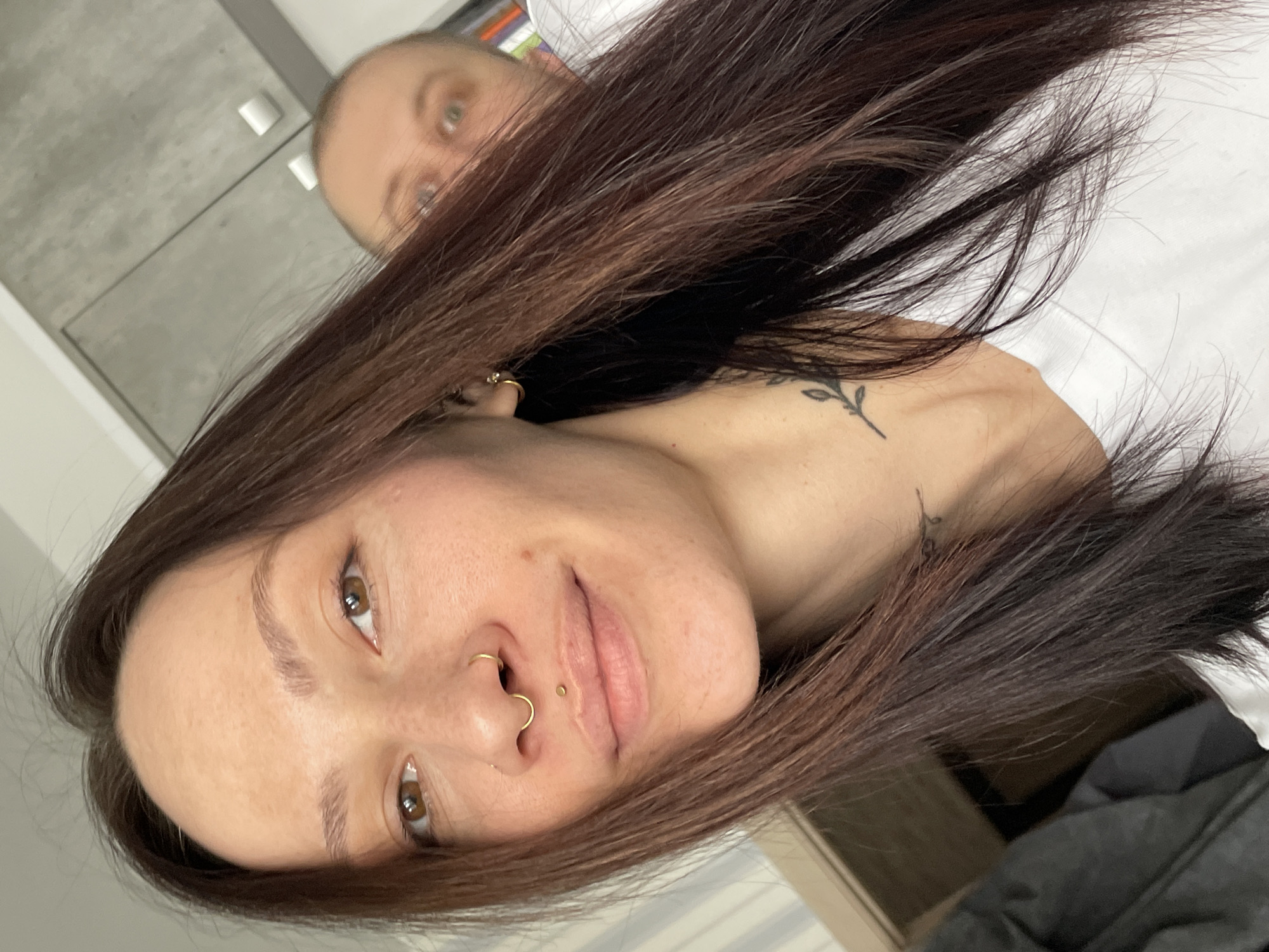 A tilted selfie depicts a woman with brown hair and facial piercings (nostril and lip). A small floral tattoo is visible on her upper chest. The background is blurred, with a partially visible face of a bald person behind her, and a cabinet in the background. There is no information to determine location in the city of Izhevsk, Russia.