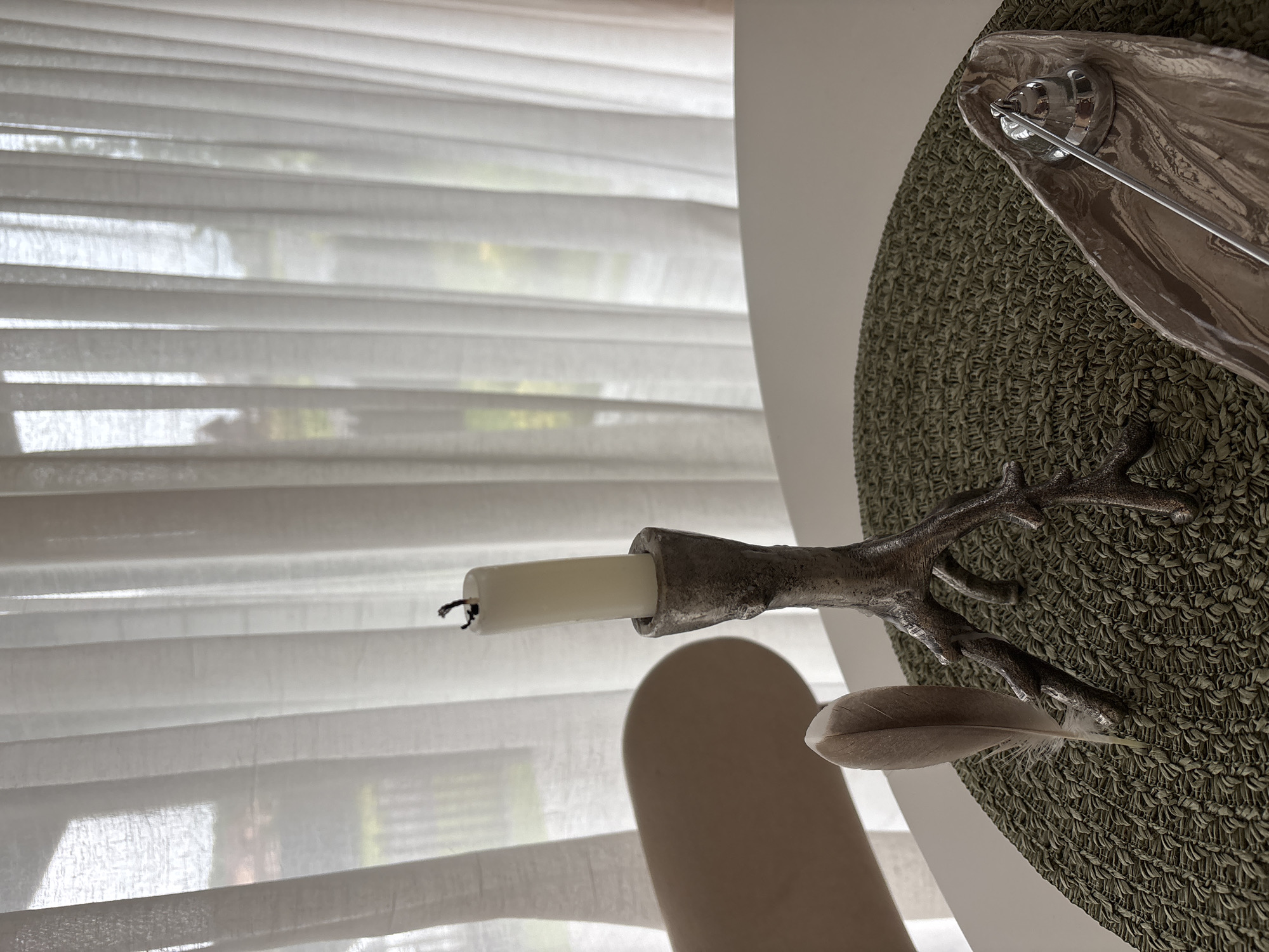 The image shows a close-up view of a table setting with decorative items. A white, unlit candle sits in a dark, branch-like candlestick. Next to the candlestick, a single feather rests on a green woven placemat. In the foreground and to the right, a decorative dish with a silver snuffer is visible. In the background, sheer white curtains are drawn, obscuring a view of what appears to be a window and possibly some greenery outside. The overall scene is indoors, with soft lighting. The location is not specified as Zvenigorod, Russia.