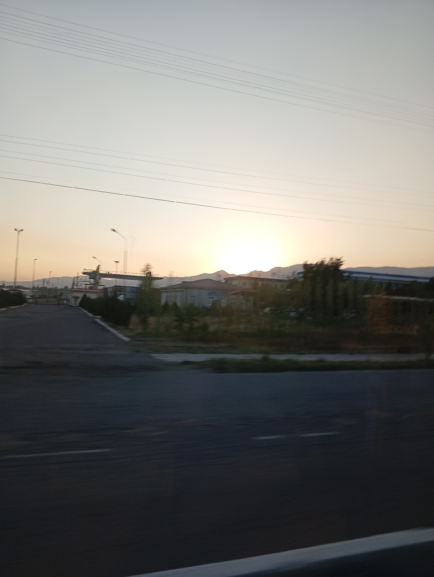 The image captures a landscape scene at dusk in Angren, Uzbekistan. The foreground is dominated by a blurred asphalt road, indicating movement, likely from a vehicle. In the middle ground, a road leads towards an industrial area characterized by buildings, fences, and overhead cranes. The setting sun is visible on the horizon, casting a warm, hazy glow and silhouetting distant hills. Power lines crisscross the sky, suggesting an urban or semi-urban environment. The overall impression is that of an industrial zone on the outskirts of Angren during sunset.