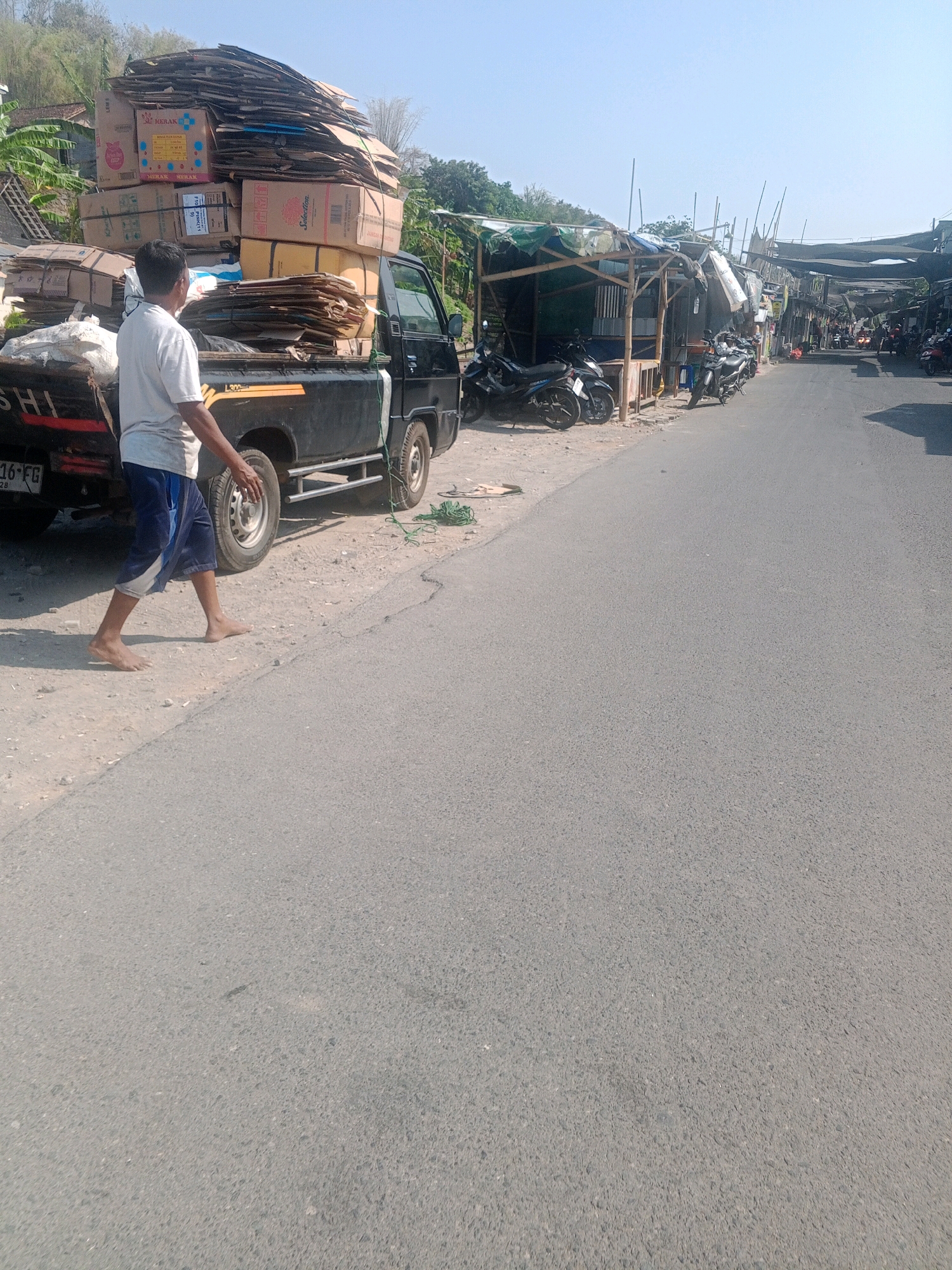 A man walks barefoot beside a black pickup truck loaded with cardboard boxes. The truck is parked on the side of a paved road in Wonogiri, Indonesia. In the background, there are several makeshift stalls with awnings and parked motorcycles, suggesting a market or roadside business area. The environment is sunny with a clear blue sky.