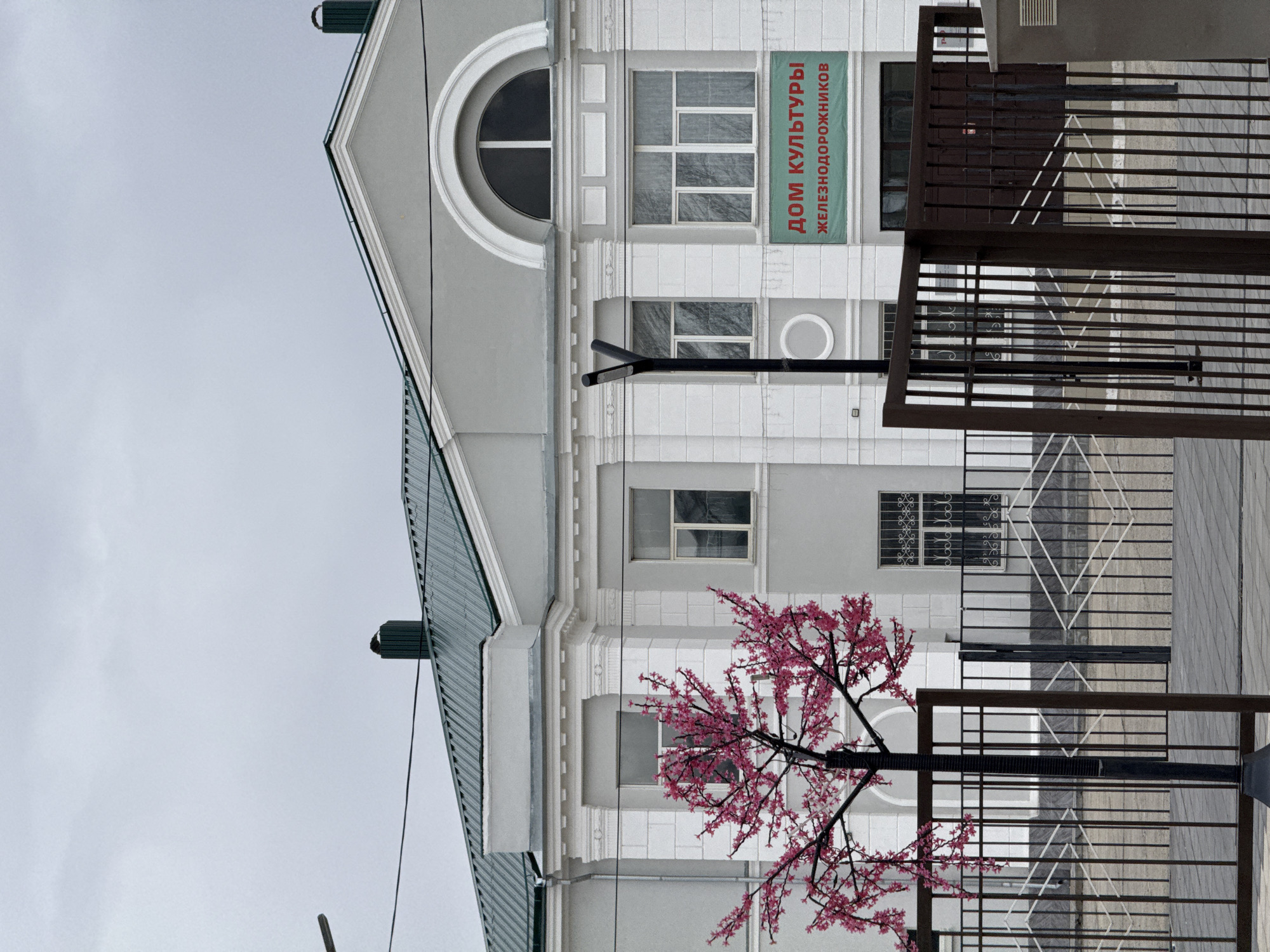 The image depicts a building in Borzya, Russia. The building is identified by a sign in Russian that translates to "House of Culture of Railroad Workers." The architectural style includes a rounded window at the top, rectangular windows, and a light gray and white color scheme. A metallic green roof is visible. In front of the building, a decorative tree with pink blossoms is situated behind a metal fence. The sky is overcast.