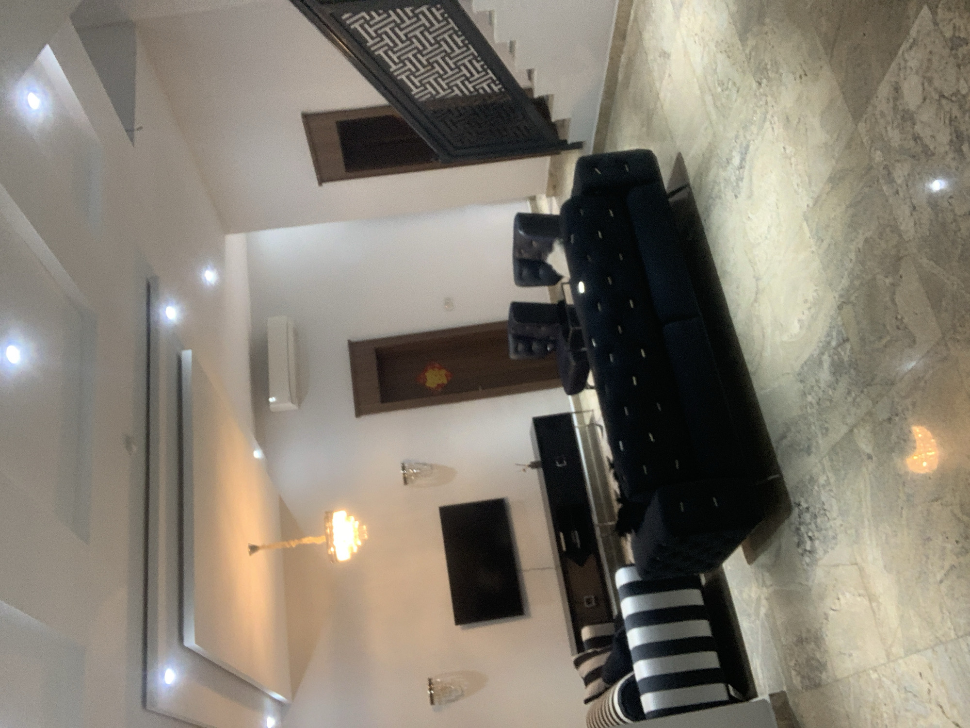 The image shows an interior view of a modern living room. The room features a high ceiling with recessed lighting and a decorative chandelier. A black sectional sofa and two black armchairs are arranged in a seating area. In the background, a black media console holds a flat-screen television. A staircase with a patterned railing is visible on the right side of the image. The floor is tiled with a beige and grey marble pattern. There are no people visible in the image. The overall impression is one of contemporary elegance and spaciousness.