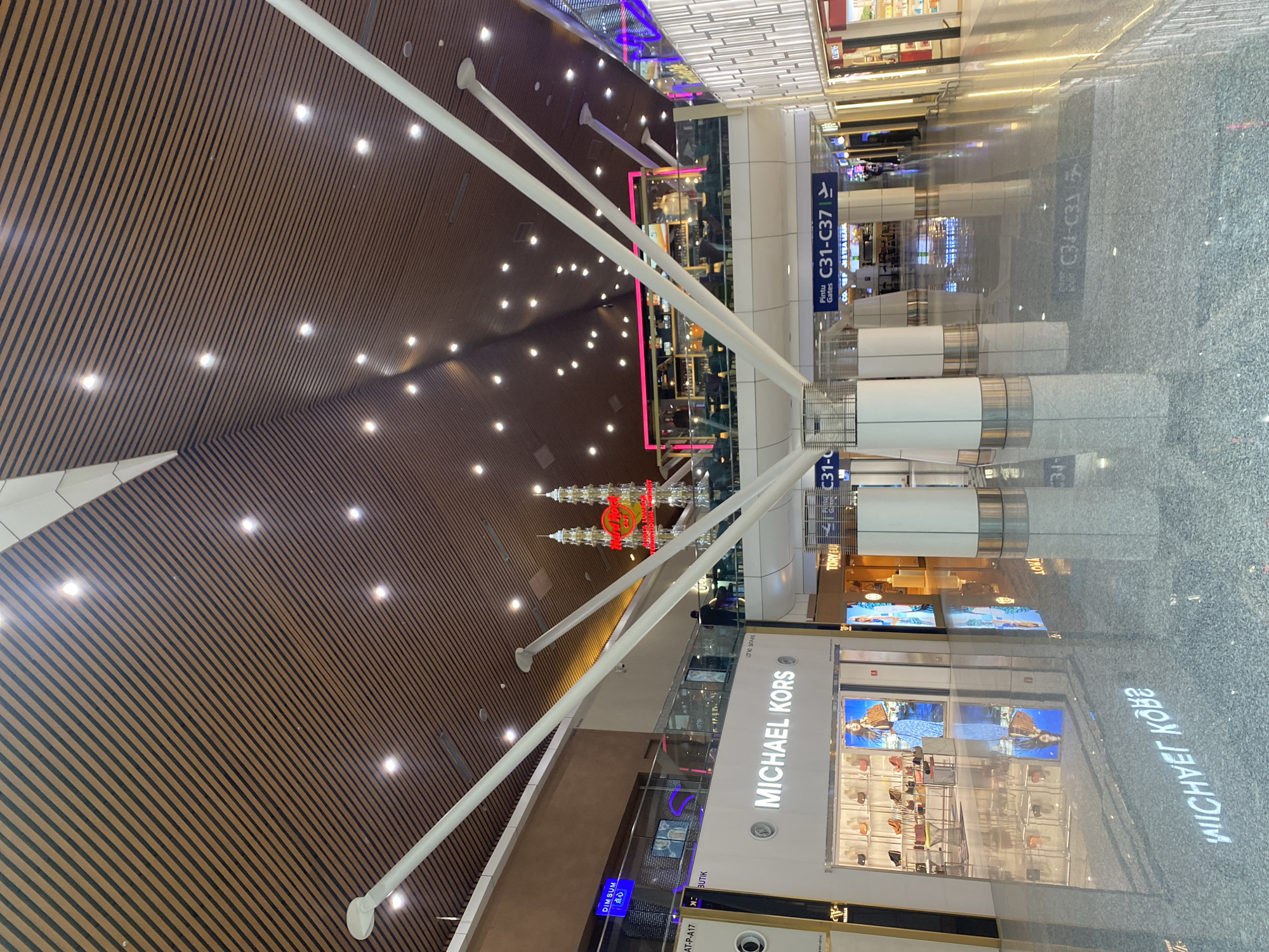 The image depicts the interior of a modern airport terminal in Sepang, Malaysia. The scene is characterized by a high, wood-paneled ceiling with integrated lighting, and a polished floor that reflects the surrounding environment. Prominently featured is a Michael Kors store with its illuminated signage and window displays showcasing handbags and a model. Adjacent to it, a Tory Burch store is visible. In the background, a Hard Rock Cafe structure with red neon signage shaped like twin towers stands out. Several white, angled structural supports extend from the ceiling, converging near the center of the frame. Directional signage indicates "Pintu Gates C31-C37." The overall environment suggests a busy commercial area within an international airport, with people visible in the distance, appearing to be moving through the terminal.