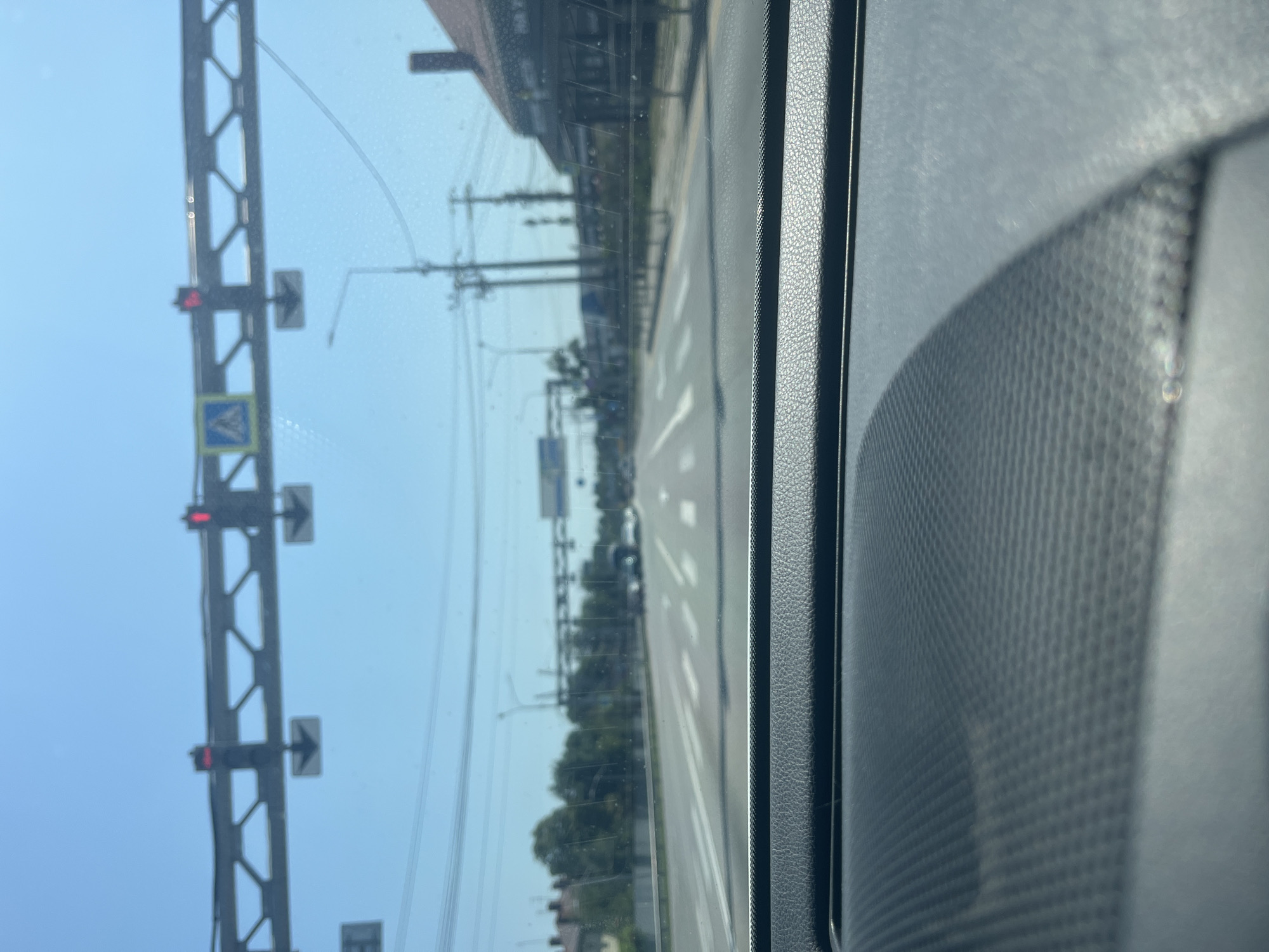 The image depicts a view from inside a vehicle looking forward on a sunny day. The perspective is from the driver's seat, showing the dashboard in the foreground. Ahead, a multi-lane road stretches into the distance, with clear lane markings and a crosswalk visible. Traffic lights are mounted on an overhead gantry, with red lights illuminated, indicating a stop. A pedestrian crossing sign is also present. Power lines and utility poles line the side of the road, with buildings and trees forming the background on either side. The sky is clear and blue.