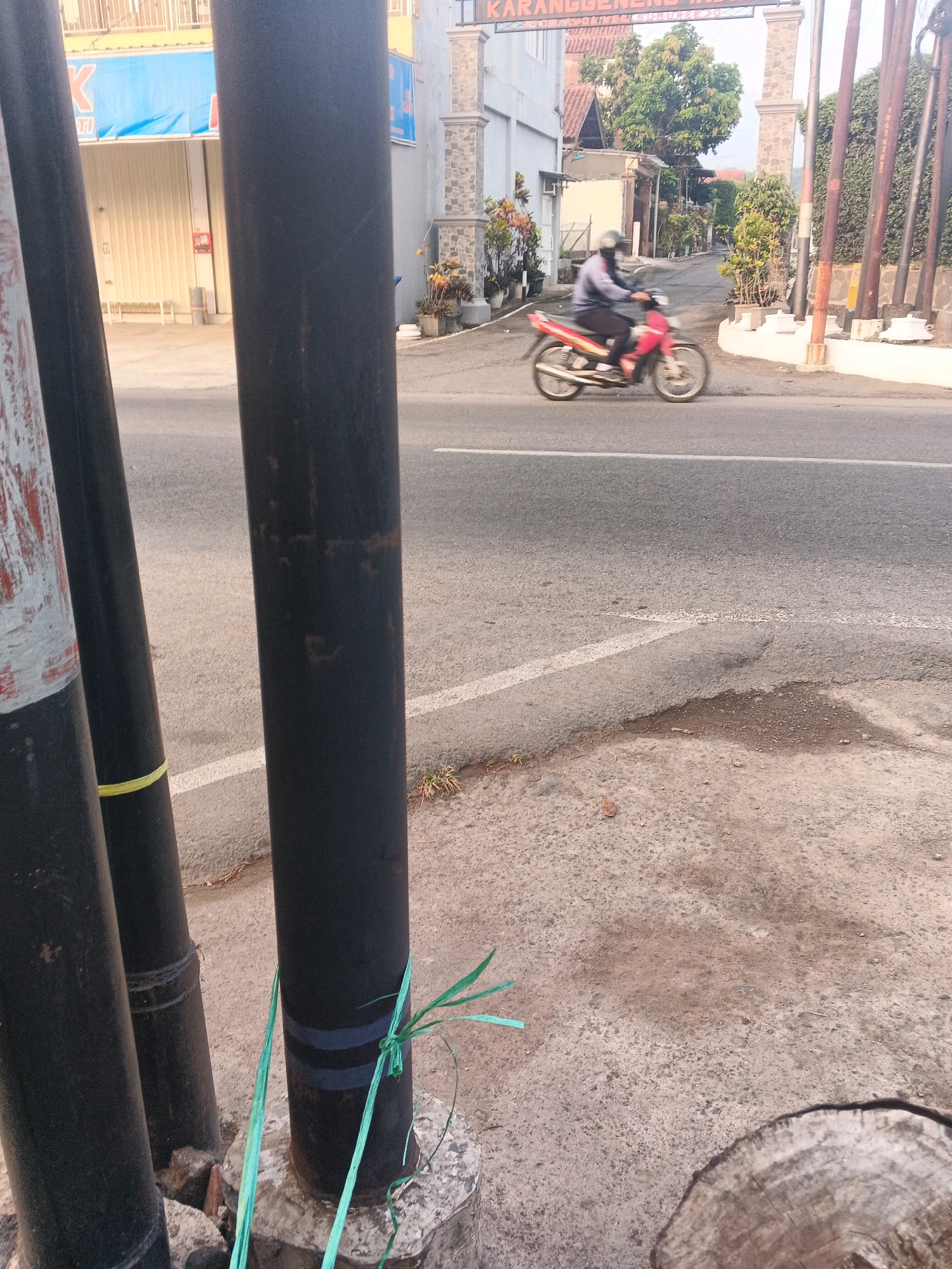 This image captures a street scene in the city of Semarang, Indonesia, likely near an entrance or gateway as suggested by the archway visible in the background with the inscription "KARANGGENENG INDAH." The foreground is dominated by several large, dark, cylindrical poles, one of which is partially wrapped with green plastic tie material. To the left, the edge of another pole with red and white markings is visible.
The middle ground shows a paved road with a white lane marker. A motorcycle rider, moving from left to right, is blurred due to motion, indicating they are traveling at a moderate speed. The rider is wearing a helmet, a light-colored long-sleeved shirt, and dark pants. The motorcycle itself is red and white.
The background reveals a street lined with buildings and greenery. On the left, a building with a blue banner and a closed roller shutter is visible, possibly a storefront. Further back, residential buildings with tiled roofs and trees are present. The overall environment suggests an urban or semi-urban setting in Indonesia during daylight hours, with warm lighting indicating either morning or late afternoon. The road surface appears to be asphalt.