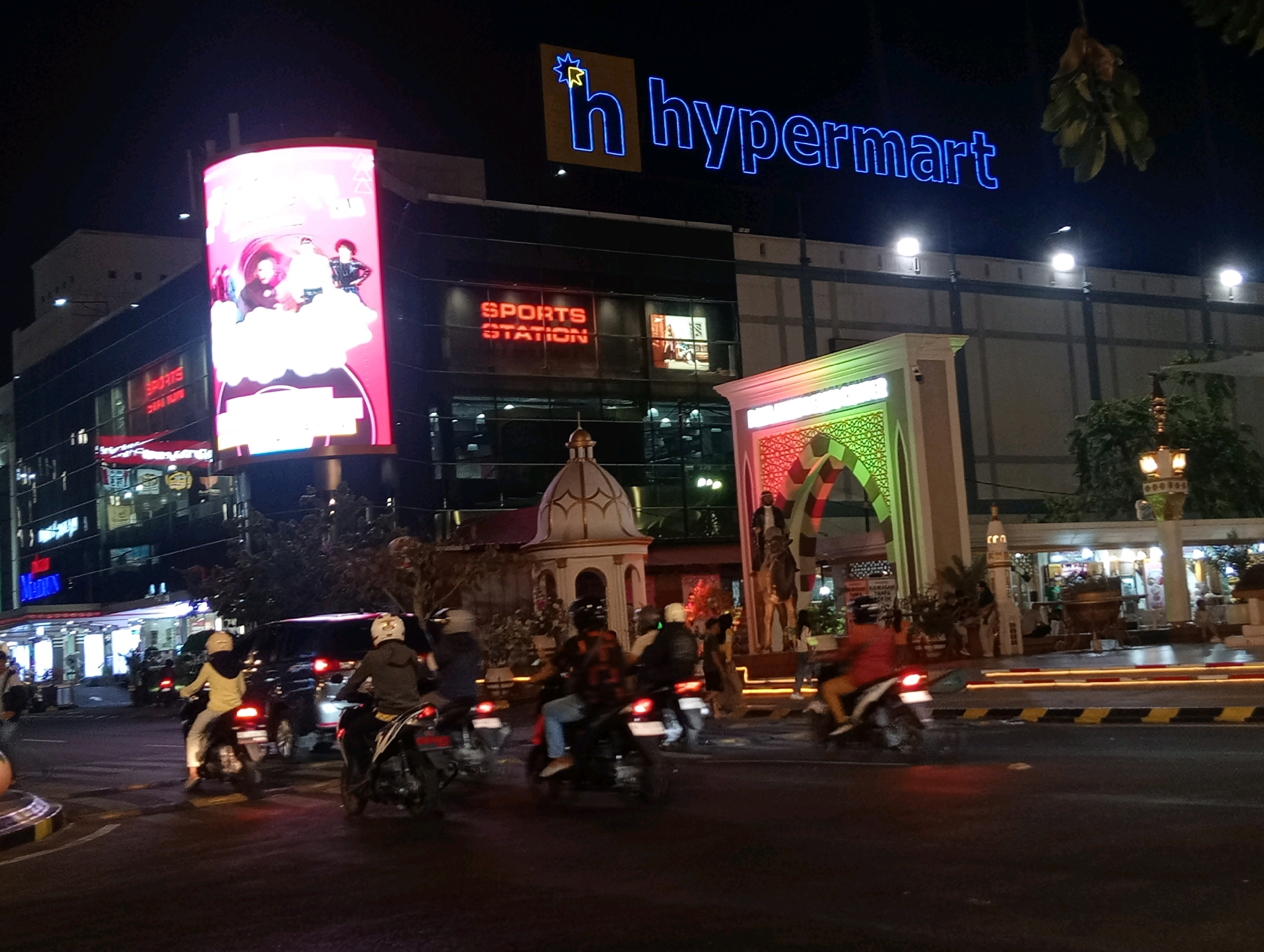 This is a night-time, eye-level, medium shot of a street scene in front of a Hypermart in Madiun, Indonesia. The Hypermart building is lit up with a large, illuminated sign that reads "h hypermart" in blue neon letters. In front of the building, there are various structures, including a dome-shaped building with intricate detailing and an arched gateway adorned with green and red patterns. Several people are riding motorcycles on the street in the foreground, many wearing helmets and casual clothing. The road is wet, suggesting recent rain, and illuminated by streetlights and the lights from the businesses. The overall atmosphere is busy and illuminated, characteristic of a commercial area at night.