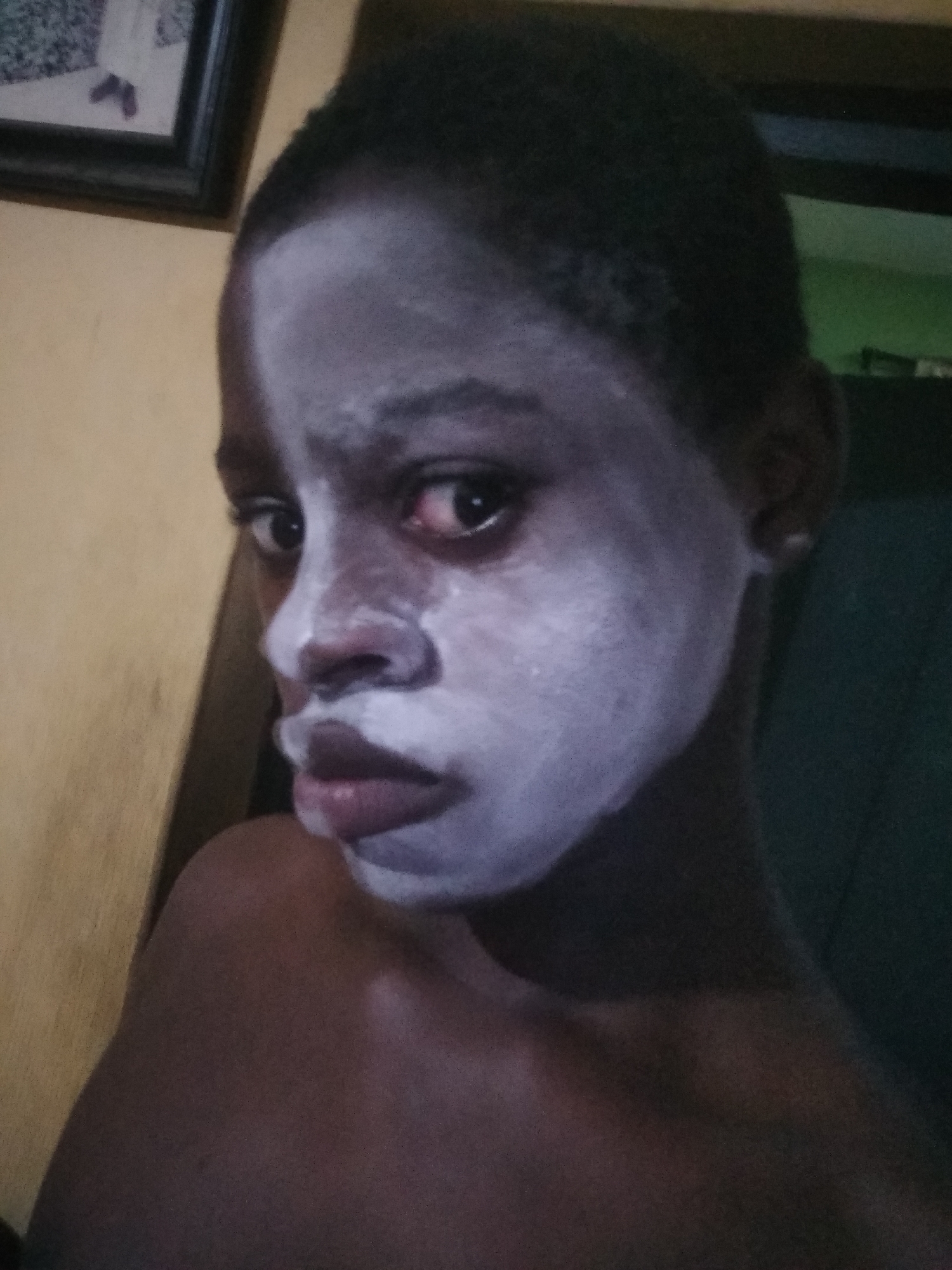 A close-up portrait of a dark-skinned child with a light-colored substance applied to their face, likely a skincare product or mask. The child has short, dark hair and is looking towards the left of the frame. Their expression appears serious or contemplative. The background is blurred but suggests an interior setting with a picture frame and what appears to be a wall or curtain. The location, Shangisha, Nigeria, is based on external information and cannot be verified solely from the image.