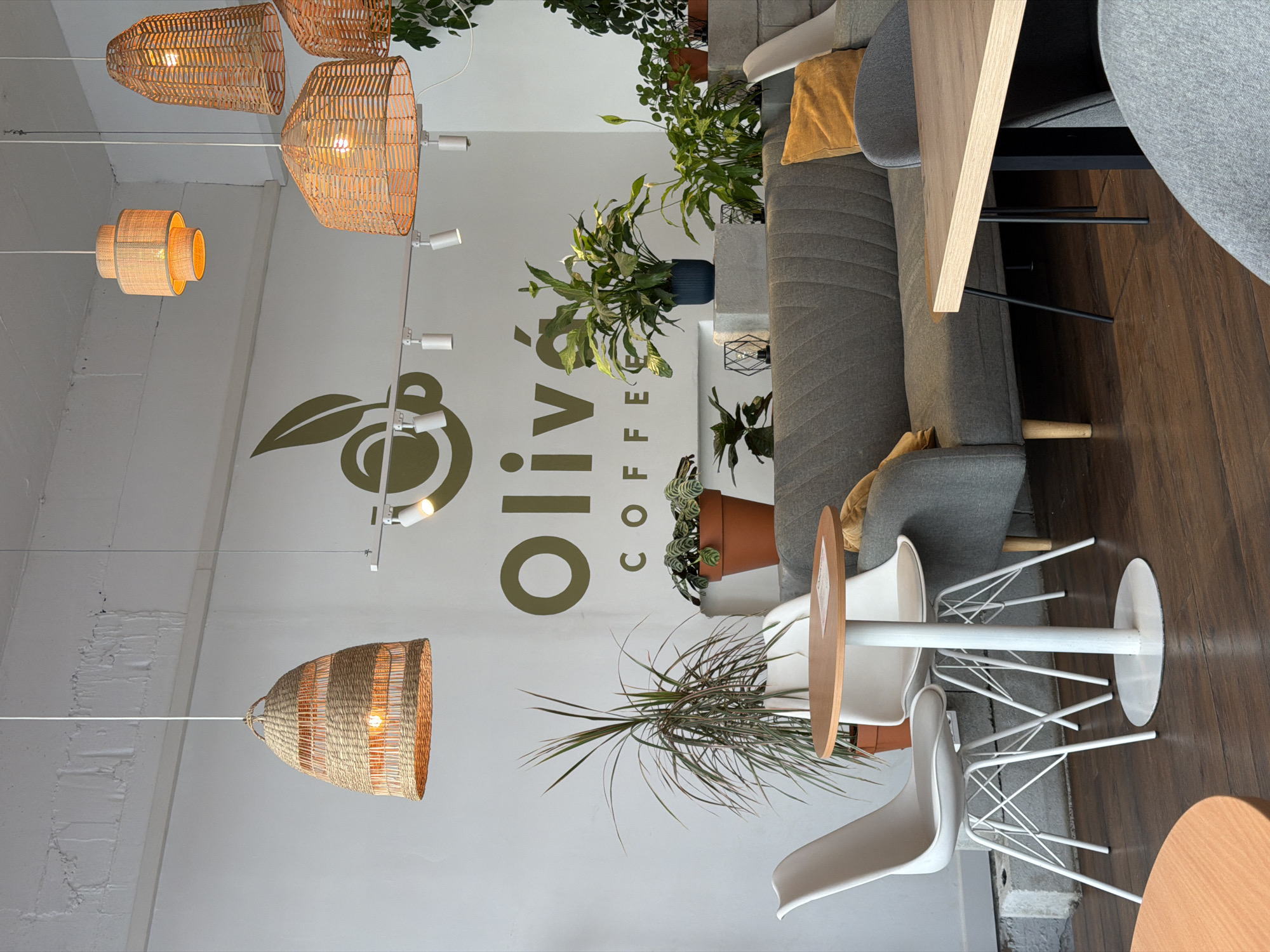The image depicts the interior of "Olivia Coffee" in Podgorica, Montenegro. The coffee shop features a modern, minimalist design with white walls and exposed brick accents. Woven pendant lights hang from the ceiling, casting a warm glow over the space. Seating includes a grey sofa with orange accent pillows and white Eames-style chairs around a circular wooden table. Several potted plants are strategically placed throughout the cafe, adding a touch of nature. The "Olivia Coffee" logo is visible on the wall. The overall ambiance suggests a relaxed and inviting atmosphere.