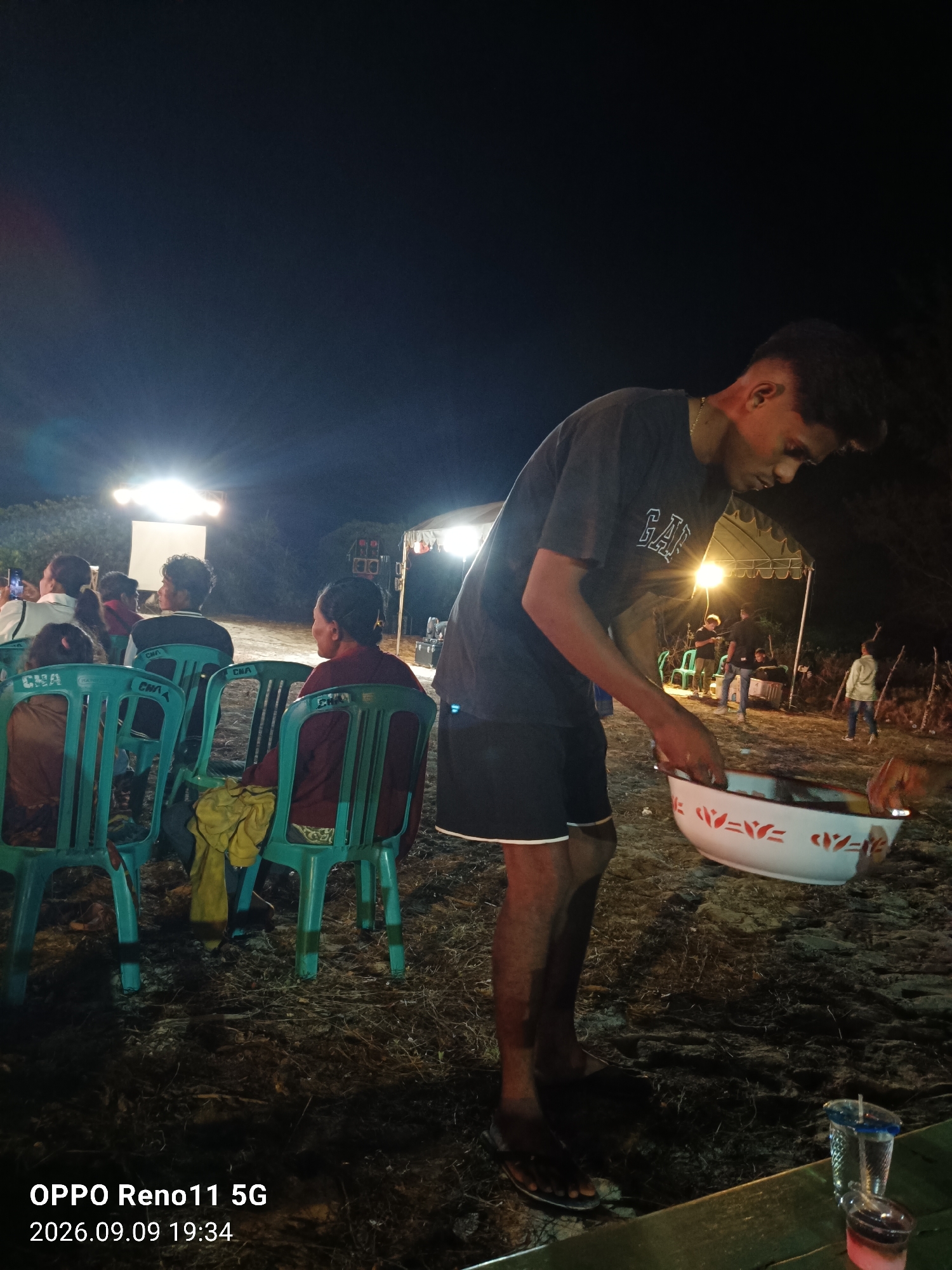 This image depicts an outdoor gathering at night in what appears to be a sandy or dirt area, likely in Otan, Indonesia, given the timestamp. The scene is illuminated by artificial lights, creating a warm glow and casting shadows.
In the foreground, a young man is prominently featured, wearing a dark t-shirt with "GAP" logo and dark shorts. He is holding a large white bowl with red decorative patterns, looking down into it. His posture suggests he is either preparing or serving something from the bowl.
In the background, several people are seated on bright teal plastic chairs, facing away from the camera. They appear to be watching something, possibly a screen or performance, as suggested by a large white screen or projector setup further in the background. Another tent structure with lighting and audio equipment is visible to the right. The environment seems to be a communal gathering space, possibly a village square or event ground. The overall atmosphere is one of casual social interaction during an evening event.