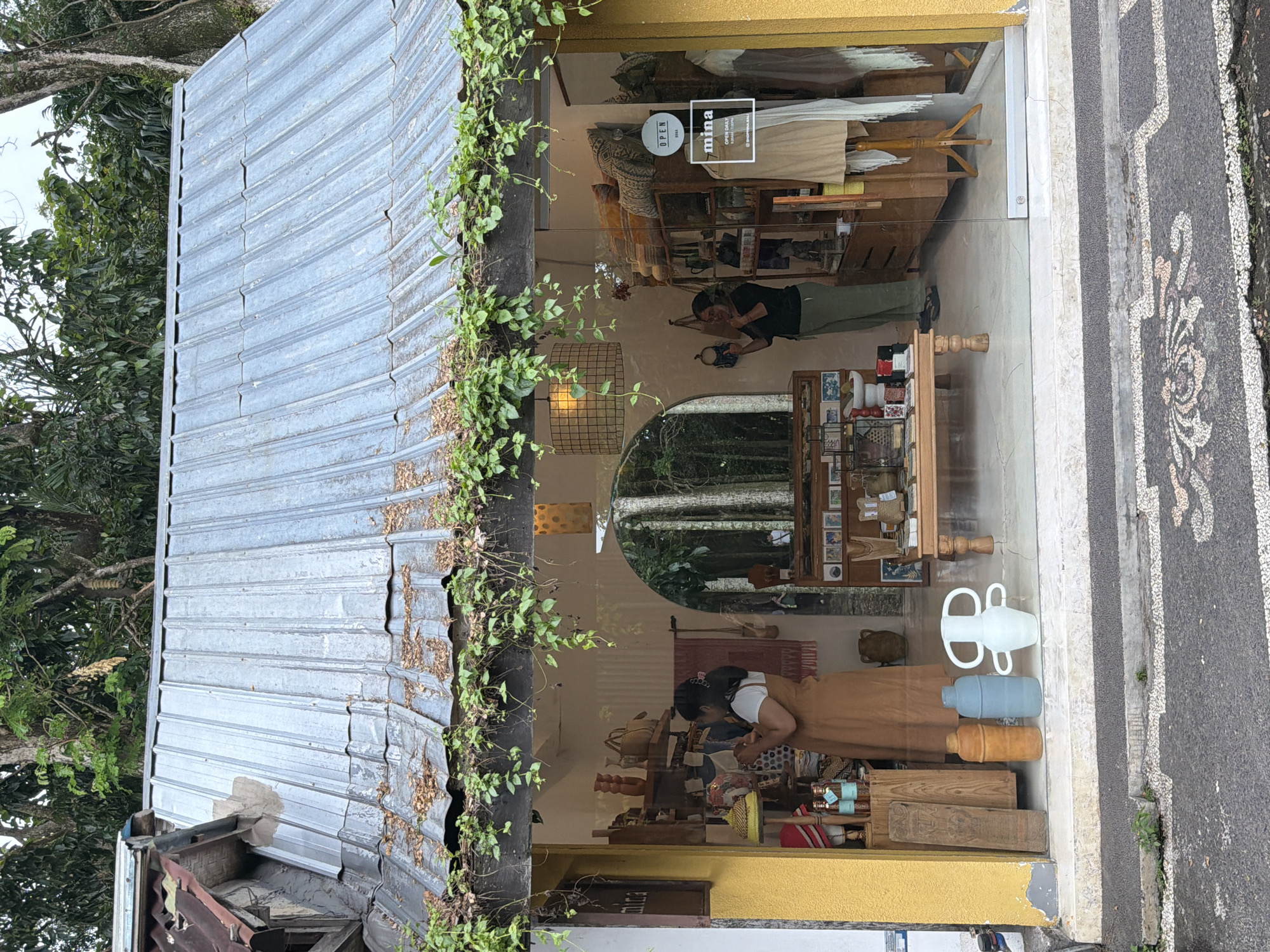 This image captures the facade of a small retail shop, likely in the Ubud District of Indonesia, viewed from the street. The shop features a prominent corrugated metal roof, partially covered in vibrant green vines that cascade down the side of the building. The front of the shop is almost entirely a large glass window, offering a view into the interior and creating reflections of the surrounding lush greenery and trees.
Inside the shop, two individuals are visible. One person, wearing a dark top and light pants, stands deeper within the store, appearing to be examining items on a wooden shelf. Closer to the storefront, another person, with dark hair and wearing a white top and a long tan/brown garment, is bent over a display table, seemingly arranging merchandise. The shop's interior is furnished with wooden shelving and display units, showcasing various items including clothing on mannequins, woven goods, small jars, bottles, and decorative objects. A distinctive round mirror on the back wall reflects more of the dense trees outside.
On the glass window, two signs are visible: a circular "OPEN" sign and a rectangular sign with the brand name "mina," along with "OPEN DAILY" and social media handles like "@SHOP.MINA.BALI" and "@MINA.BALI". On the immediate exterior, to the right of the shop, a patterned grey and black sidewalk or pathway adds a textural element. The lighting suggests it is daytime, possibly overcast or in a shaded area, as there are no harsh shadows. Three decorative vessels – a white pitcher-like object, a light blue cylindrical bottle, and a tan cylindrical bottle – are displayed prominently near the bottom of the window, partially obscuring the view of the pavement.