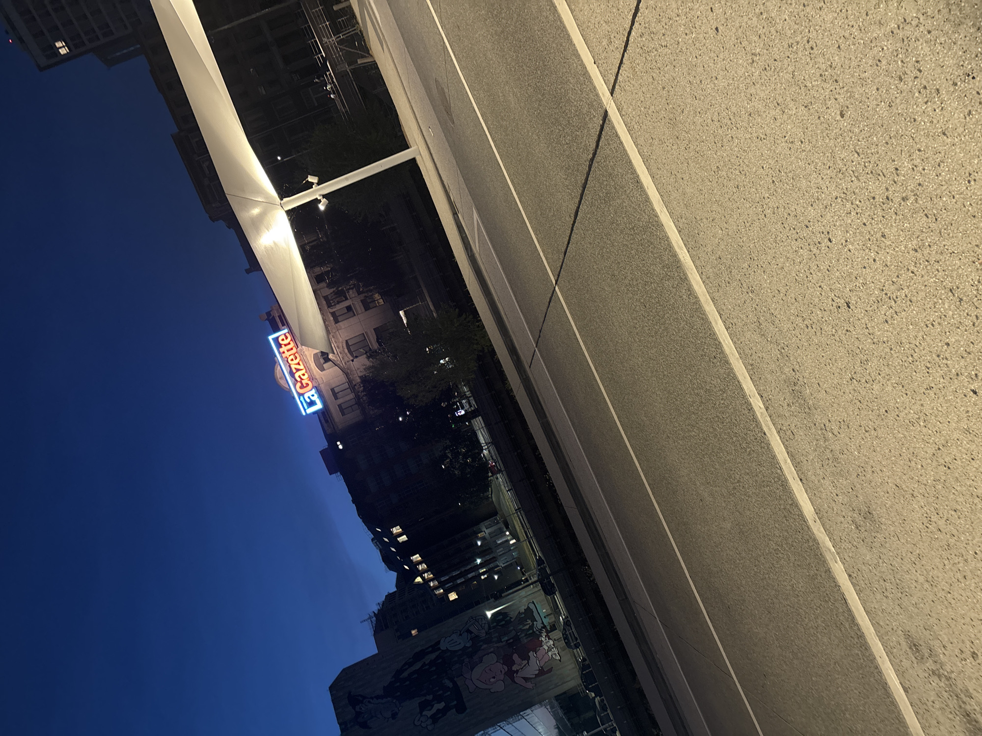 The image depicts a nighttime scene in Charleroi, Belgium, focusing on a street with a prominent mural on a building to the left. The mural features stylized cartoon characters, including what appears to be a large, dark figure and smaller figures of a girl and cartoon characters resembling Mickey Mouse. Above and to the right of the mural, a building displays a neon sign reading "La Gazette" in blue and red letters. A large, white, tent-like structure is illuminated by lights and supported by a pole, casting a shadow across the street. The street itself is paved and has white lane markings. In the background, other buildings, trees, and a railing are visible. The sky is a deep blue, characteristic of twilight or nighttime. There are no identifiable people in the foreground or midground.