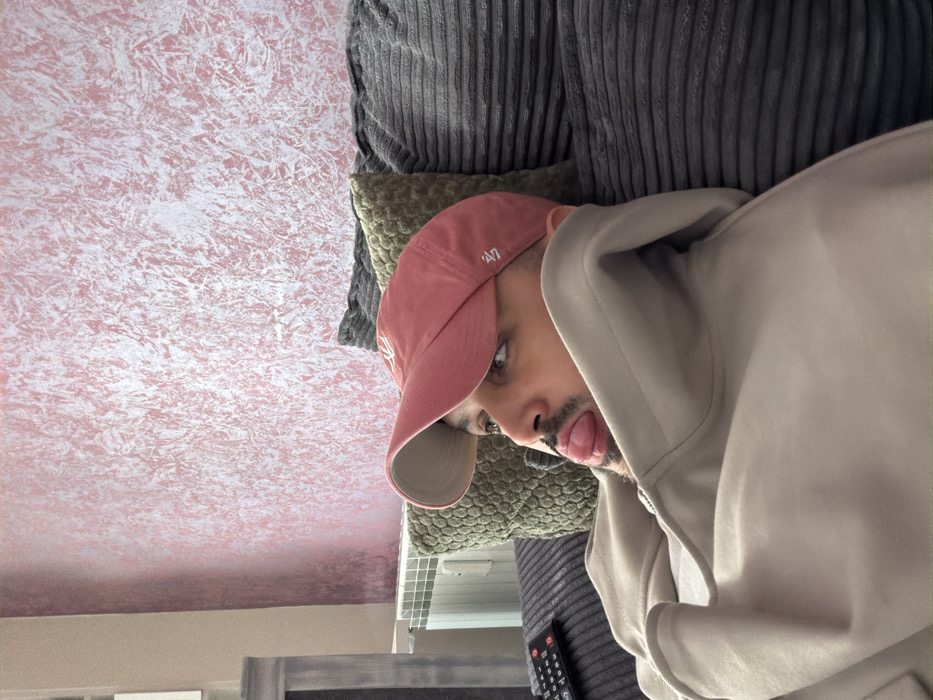 An adult male is depicted reclining on a dark grey, ribbed sofa within an indoor setting. He wears a light beige hooded sweatshirt and a faded reddish-pink baseball cap with a white "47" logo embroidered on the front. His head is turned to the viewer's right, with his right eye visible, and his gaze is directed upwards and to the right. His tongue is extended from his mouth. A short beard or stubble is present on his face. Behind his head, an olive-green, textured square pillow is visible, supported by a dark grey, ribbed pillow. To the left of the individual, a black remote control with red buttons rests on the sofa cushion. The background wall features a textured decorative finish with a base color of reddish-pink and irregular white highlights. A white horizontal panel radiator is installed along the lower left wall. A segment of a grey furniture piece is partially visible on the far left. The specific location within Sofia, Bulgaria, cannot be determined from the visual information provided in the image.