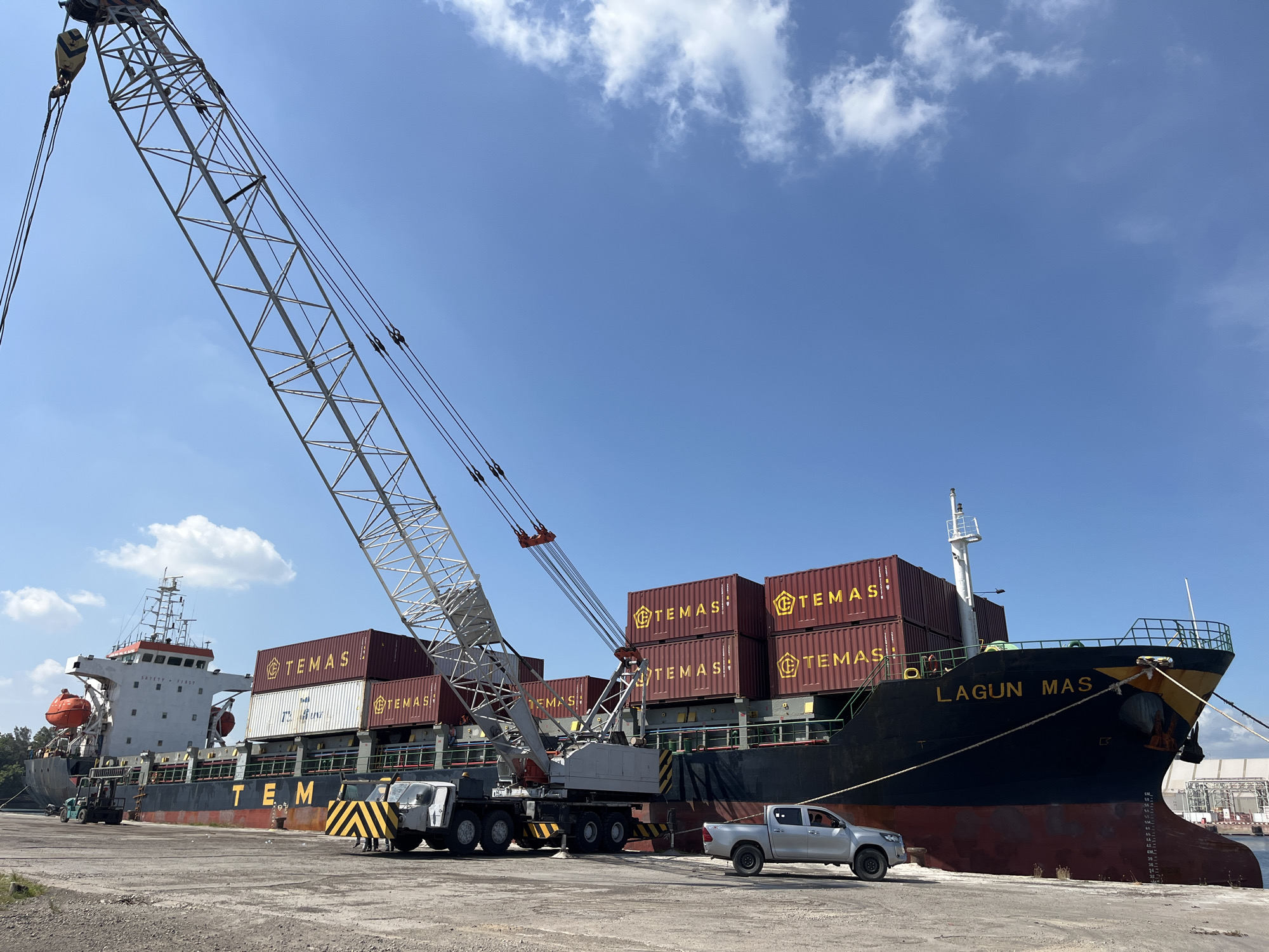 The image depicts a cargo ship named "LAGUN MAS," marked with the word "TEMAS" on its containers, docked at a port facility in Tambonbaroh, Indonesia. A large mobile crane is positioned alongside the ship. A silver pickup truck is parked near the bow of the ship. The sky is blue with scattered clouds. The ship is partially loaded with containers.