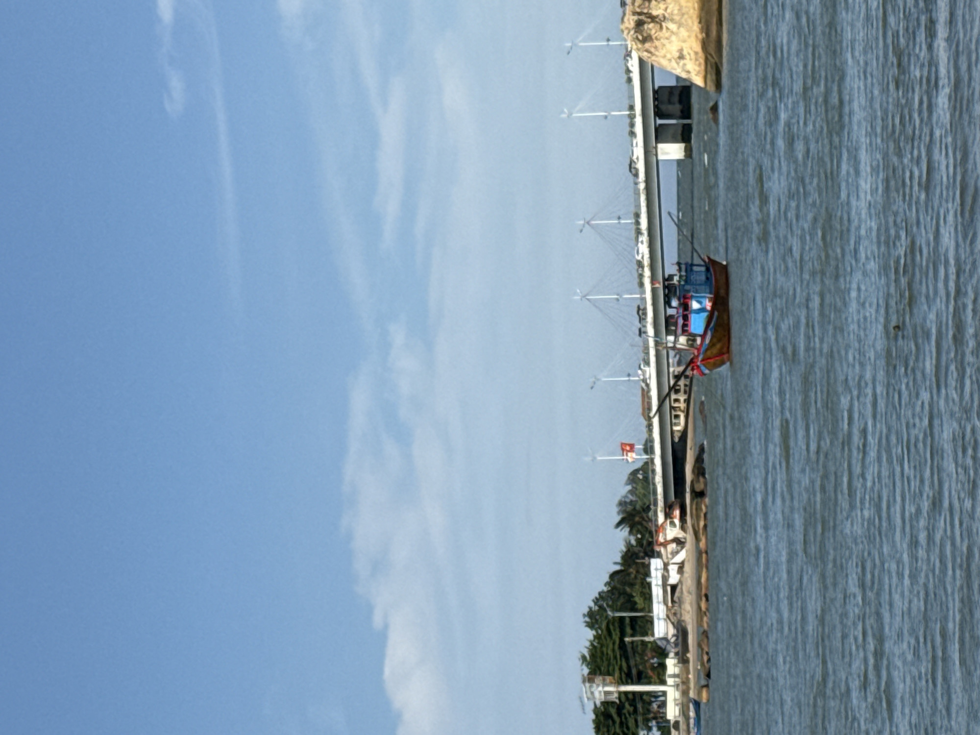 The image shows a body of water with a small fishing boat in the foreground. The boat is colorful, with red, blue, and yellow accents. In the background, a long bridge spans the water, with cars visible on it. There are also trees and buildings on the shore to the left, and a large rock formation to the right. The sky is blue with some scattered clouds. The scene appears to be a coastal area in Bac Nha Trang Ward, Vietnam.