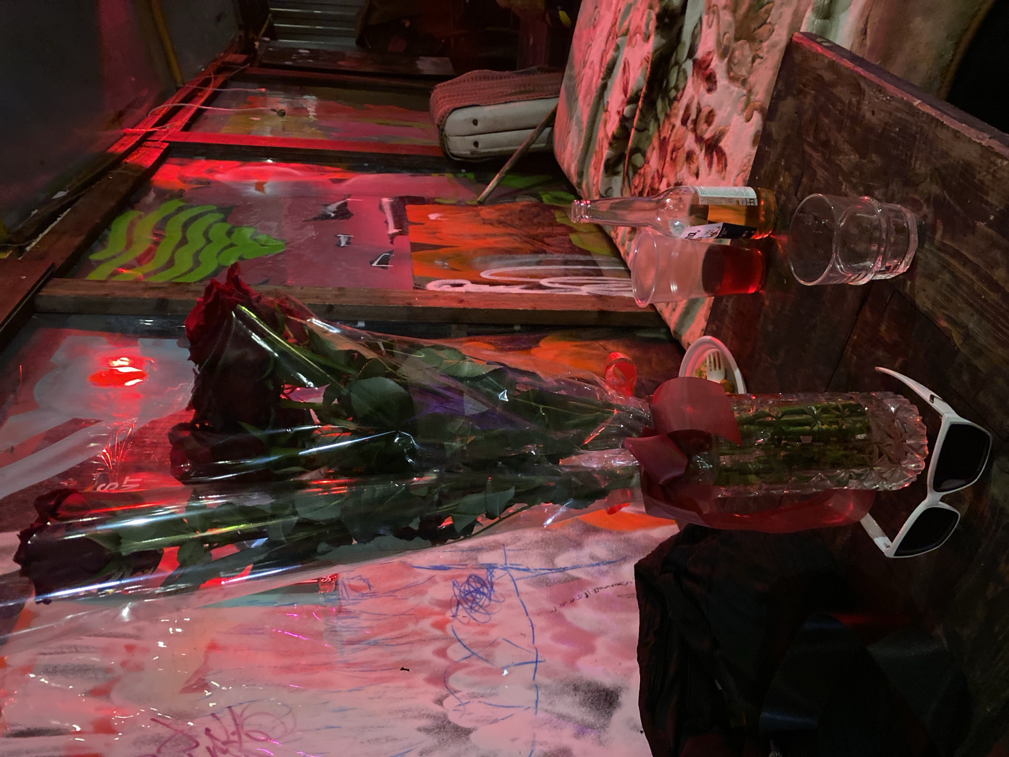 The image shows a cluttered surface with various items, possibly located in a city, potentially Moscow, Russia. Key objects include a bouquet of dark roses wrapped in clear plastic, a crystal vase with greenery tied with a red bow, a pair of white sunglasses, two clear drinking glasses, one with red liquid, and an empty bottle. The surface beneath these items appears to be decorated with graffiti and drawings in pink, purple, and blue. Additionally, a cushion and a piece of furniture are visible in the background. The scene is illuminated with a red light, casting a vibrant hue across the objects and surface.