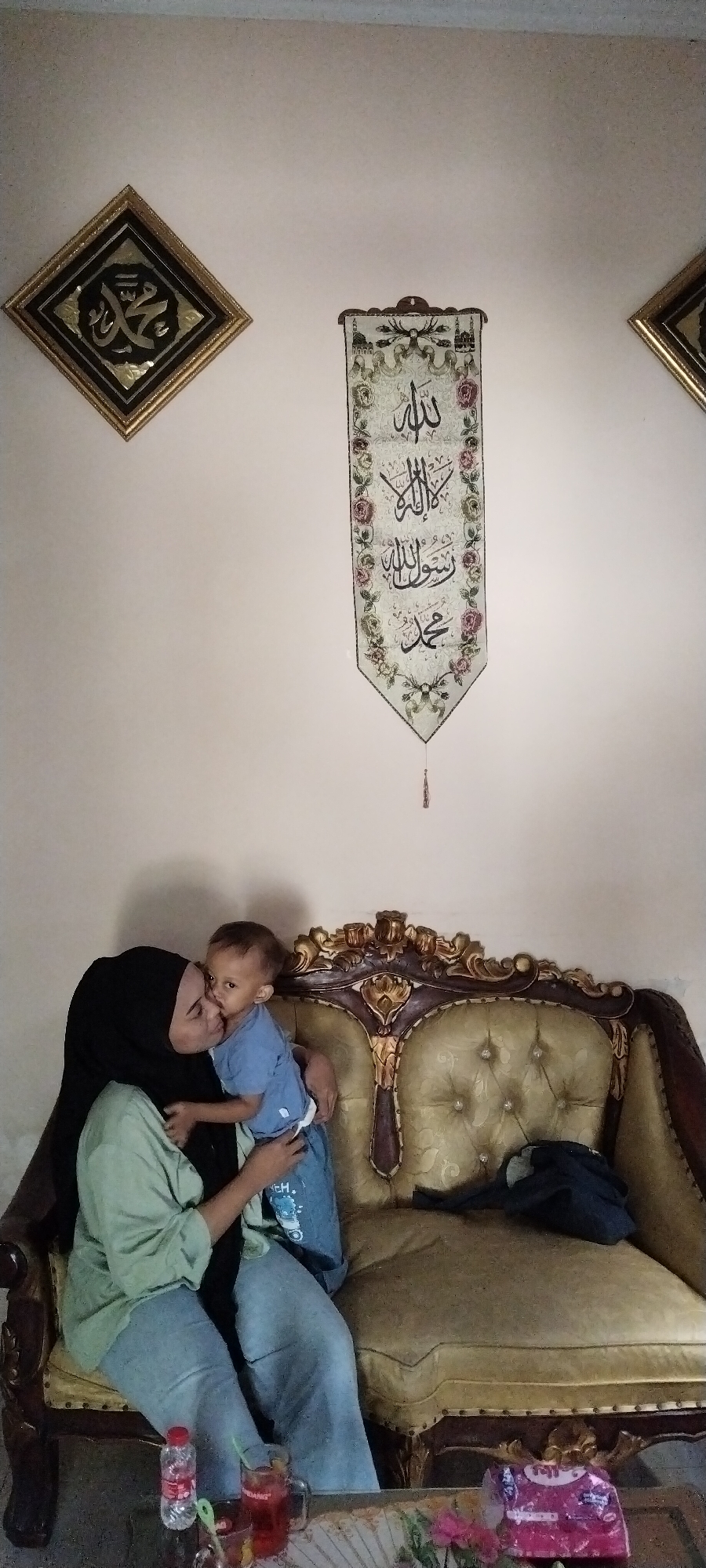 A woman wearing a black hijab and a light green shirt is sitting on a gold-accented ornate sofa, holding a young child who is wearing a blue shirt and jeans. The woman is kissing the child on the cheek. The sofa is in a room with a light-colored wall. Two framed Islamic calligraphy artworks are hung on the wall: one on the left, a square frame with a gold border and black background, and another in the center, a vertical banner with floral decorations and Arabic script. A dark blue jacket is draped over the right side of the sofa. In the foreground, on a table, are a clear plastic bottle of water, a glass mug with a red beverage, and a pink package of tissues. The scene is likely a home environment in the city of Peganjaran, Indonesia.