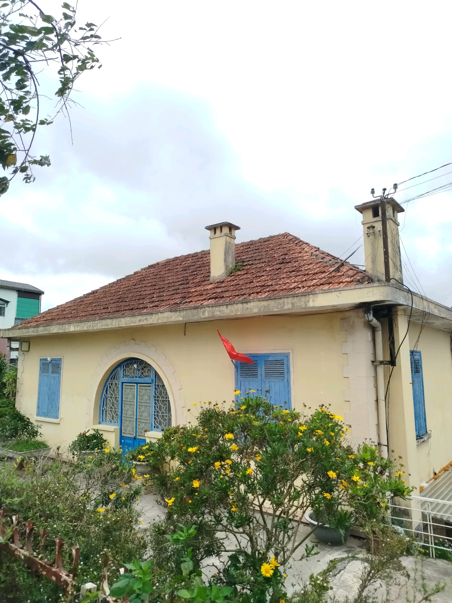 The image depicts a light yellow, single-story house with a terracotta tile roof. The house features blue shutters on its windows and a distinctive arched doorway with intricate metalwork. A small red flag is visible on the side of the house. In the foreground, lush green bushes with yellow flowers are in bloom. The house is situated in a residential area, with a glimpse of another building with green shutters visible in the background to the left. An old, rusty metal fence runs along the bottom of the frame. The sky is overcast, suggesting a cloudy day. This type of architecture and vegetation is characteristic of the charming French colonial villas found in the city of Da Lat, Vietnam, particularly in areas like the Xuan Huong Lake vicinity or the old French Quarter.
