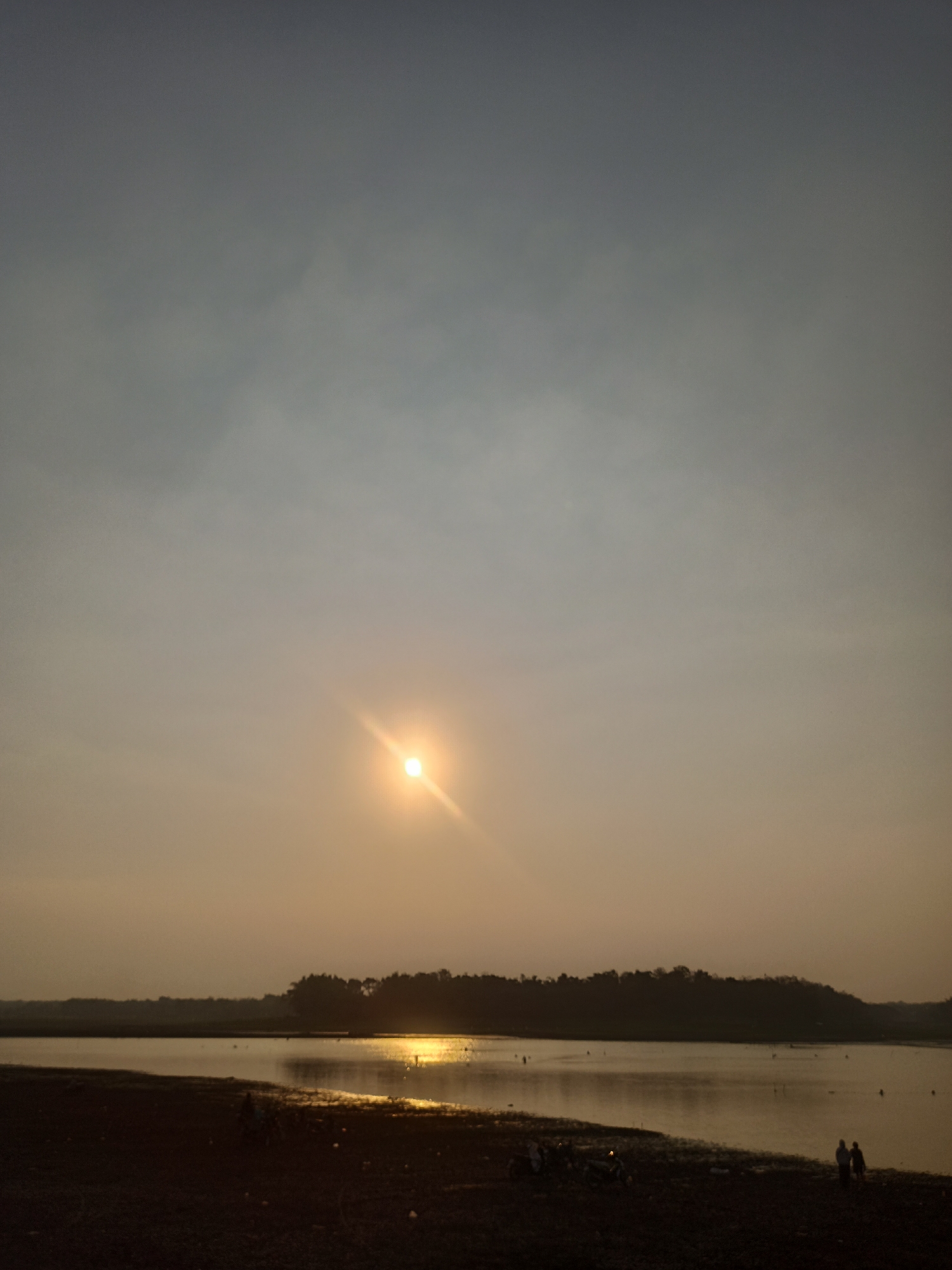 The image captures a serene sunset over a body of water, likely a lake or reservoir, with a silhouetted treeline forming the horizon. The sun, a bright orb with a visible lens flare, hangs low in the sky, casting a warm, golden light across the water's surface. The sky transitions from a pale blue at the top to a soft orange and yellow hue near the horizon, suggesting the late stages of daylight.

The foreground depicts a dark, earthy shoreline, with small pebbles and scattered debris. A few individuals are visible on the shore, with two figures standing close together on the right side, appearing to be watching the sunset. Several motorcycles are parked near the left edge of the frame, adding a touch of human presence to the otherwise natural landscape. The water itself is calm, reflecting the vibrant colors of the sky.

While the specific location within Tanon, Indonesia, is not explicitly provided, the scene evokes a sense of a peaceful, natural area, perhaps a popular spot for locals to enjoy the evening. The overall atmosphere is tranquil and reflective.