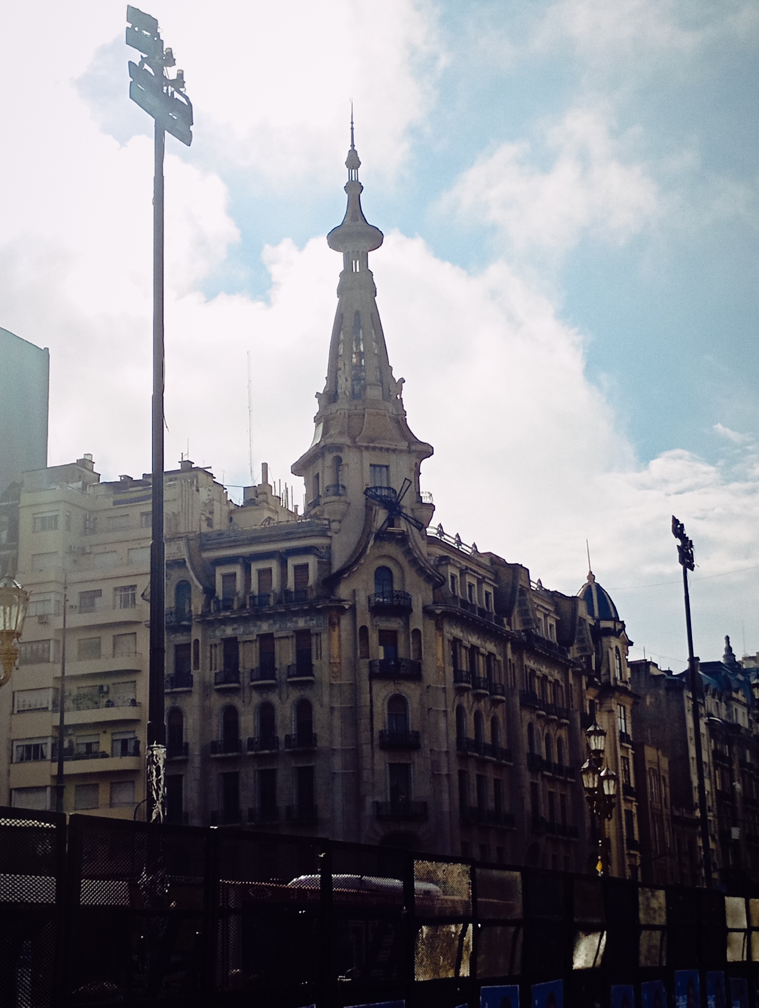 The image presents a daytime street scene in Buenos Aires, Argentina, featuring a prominent, ornate building with a distinctive steeple, likely the Palacio de la Prensa. This central, multi-story structure exhibits classical architectural details, including arched windows, decorative balconies, and a tall, elaborate spire at its apex, with smaller domes visible on adjacent sections.
To the left of the main building stands a more modern, lighter-colored apartment building. To the right, a series of older buildings with varied facades and some additional domes extend down the street. The sky is partially cloudy, showcasing patches of blue amidst white clouds, indicating clear weather.
Two tall, dark utility poles are prominent, one on the left fitted with large, rectangular floodlights or light panels, and another on the right. An antique-style, multi-lamp street light is partially visible in the lower-left foreground. A dark, mesh-like barrier or fence occupies the immediate foreground, obscuring the street level and showing reflections, including a faint outline of a red vehicle. There are no visible people or discernible activities. No legible text is present in the image.