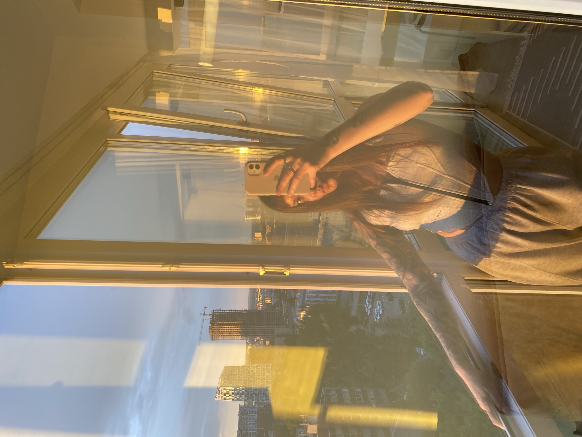 The image shows a young woman taking a selfie in a room with a large window, likely a balcony or sunroom, at sunset. The light from the setting sun casts a warm, golden glow across the scene, reflecting off the glass. The woman is wearing a light-colored top and pants. She is holding a smartphone in her hand, using it to capture her reflection. Her arm, adorned with tattoos, is visible as she holds the phone.
Through the window, a cityscape is visible, featuring buildings under construction and some that are already built. The sky is a pale blue with some clouds, characteristic of a clear sunset. The reflection on the glass also shows parts of the interior, including a rug and what appears to be a curtain or sheer fabric. The overall mood is relaxed and contemplative, with the golden hour light contributing to a peaceful atmosphere. The time of day is clearly sunset, given the warm, low-angled light and the cityscape bathed in this light. The location cues suggest an urban environment, and the provided context indicates Izhevsk, Russia. There is no visible text in the image.