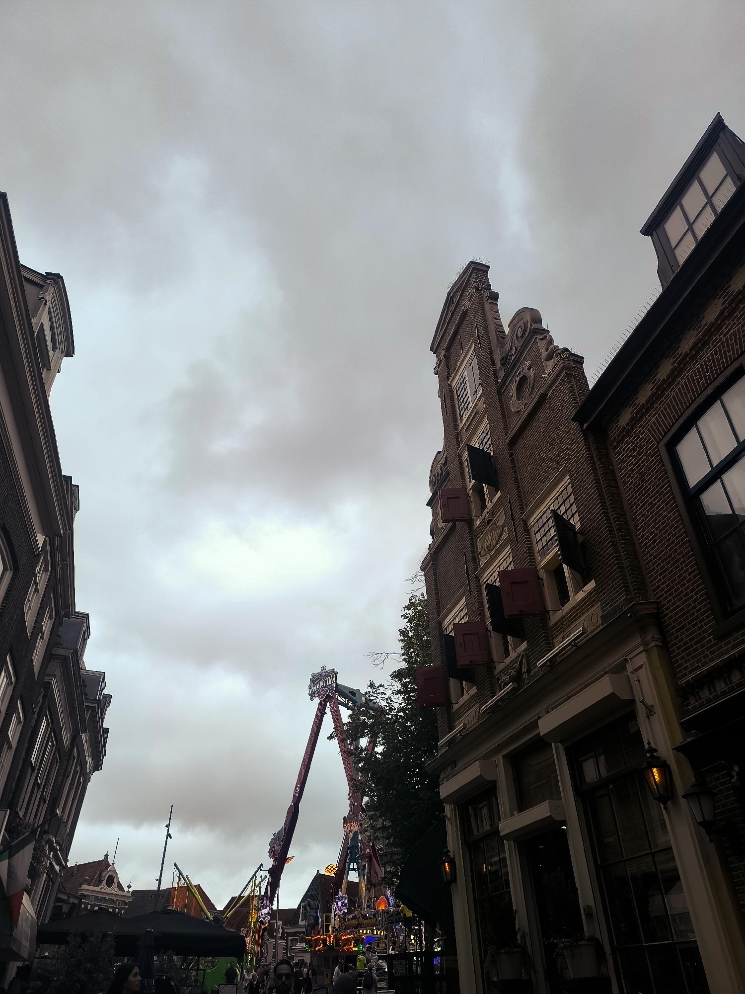The image depicts a street scene in Hoorn, Netherlands, under a cloudy sky. On the right side, a tall brick building with Dutch Renaissance architecture dominates the frame. It features multiple windows with dark shutters, some of which are a deep red. The building's facade is adorned with decorative elements typical of the period. On the left, another building, also dark-colored, is partially visible, leaning into the frame.
In the background, a large amusement park ride, identified as "Phantom" by its prominent signage, stands out against the sky. The ride features a towering structure with a swinging pendulum, suggesting it is in operation. Scattered around the ride and behind it are other carnival attractions, tents, and bright lights, indicating a festive atmosphere. People are visible in the lower part of the image, milling about in front of the buildings and near the attractions, suggesting a public event or fair. The overall impression is of a lively urban setting with a blend of historical architecture and modern entertainment.