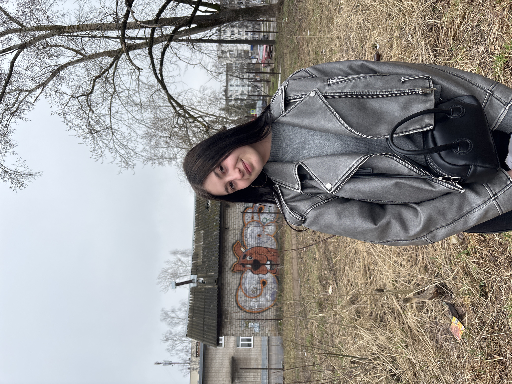 A young woman stands outdoors in Chaikovsky, Russia, near a low, gray brick building covered in graffiti of a dog character. She is smiling towards the camera. She wears a gray sweater, a gray leather jacket, and a small black handbag. The ground is covered in dry grass and twigs. Bare trees and overcast sky are visible in the background, along with residential buildings. The image is slightly rotated clockwise.
