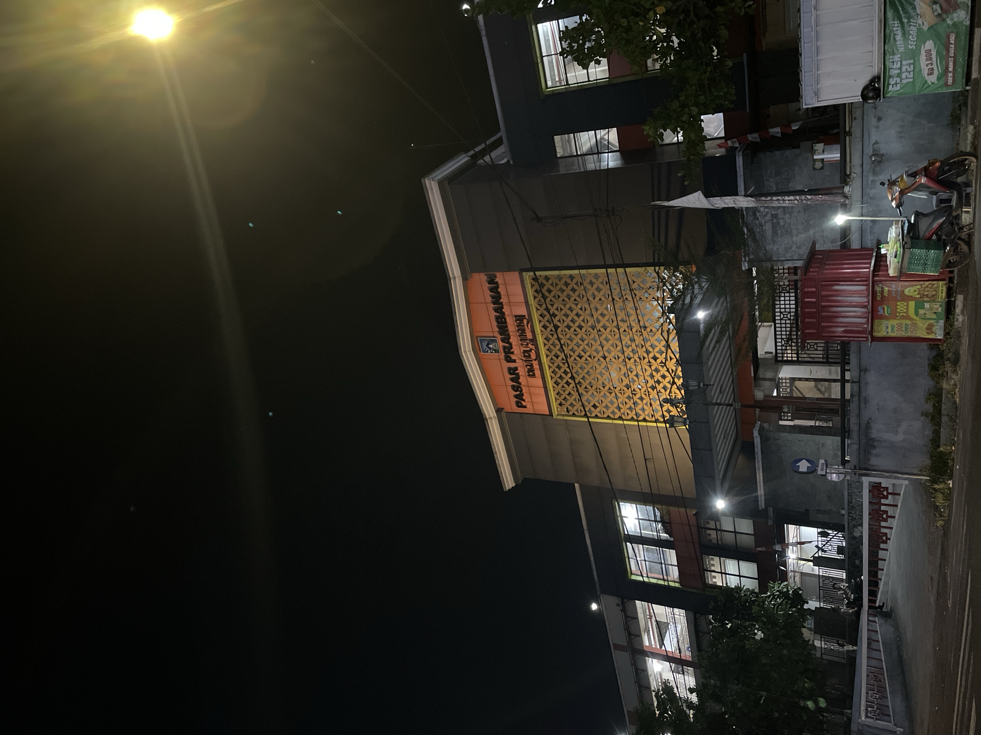 This is a nighttime, eye-level shot of the Pasar Prambanan building, illuminated by artificial lights and a bright light source in the upper right corner, possibly a streetlamp or camera flare. The building, a large, modern structure with a prominent orange facade and dark gray accents, is partially visible. Text on the orange section reads "PASAR PRAMBANAN" in large letters, with smaller text below that appears to be in Javanese script. A decorative golden lattice pattern is featured on the facade.
The lower part of the image shows a street with a paved surface. To the left, a red and white barricade is present. Further left, a tree with dark foliage is visible. In the center-left foreground, a red food cart with green shelving is parked, displaying various items and text including "Rp 3.000" and "Rp 5.000". Beside the cart is a red scooter. To the right of the food cart, another stall or structure is visible. Further to the right, a white container-like structure is present, with a green banner attached that reads "ES TEH 1221 NIKMAT!" and "Rp 3.000".
The sky is dark, with a few faint light dots, possibly stars or distant lights. The overall scene suggests a commercial area at night, with street vendors and the market building as the primary focus.