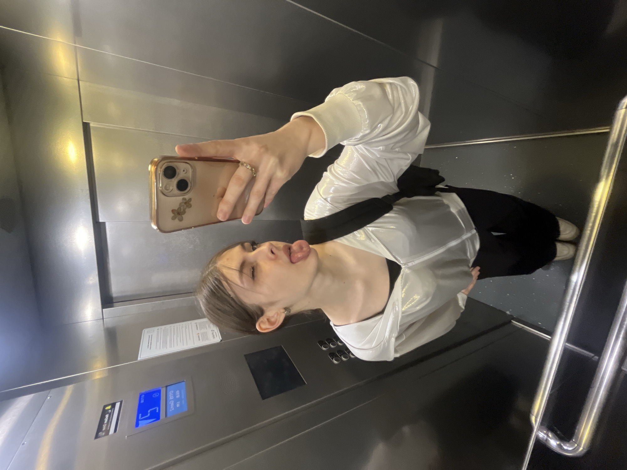 A young person is captured in a mirror selfie taken inside an elevator, rotated ninety degrees to the left. The person is holding a light gold-colored iPhone with a clear case decorated with small floral stickers in their right hand, which also features a ring on the ring finger. They are making a playful expression, with eyes closed and tongue sticking out.
The person is dressed in a shiny white long-sleeved jacket over a black top, paired with black pants and light-colored shoes. The elevator interior consists of metallic, possibly brushed steel, walls. A black handrail runs along the right side of the elevator, and the floor is dark with subtle speckles.
On the elevator control panel, a blue digital display clearly shows the Russian word "этаж" (etazh), meaning "floor," with the number "5" below it, indicating the current floor. There is also a white informational notice affixed to the wall next to the person's head, containing several lines of text in Russian, which appears to be instructions or regulations. The reflection on the phone screen shows the camera interface, but no specific text is legible there. The overall mood conveyed by the pose is one of lightheartedness and fun.