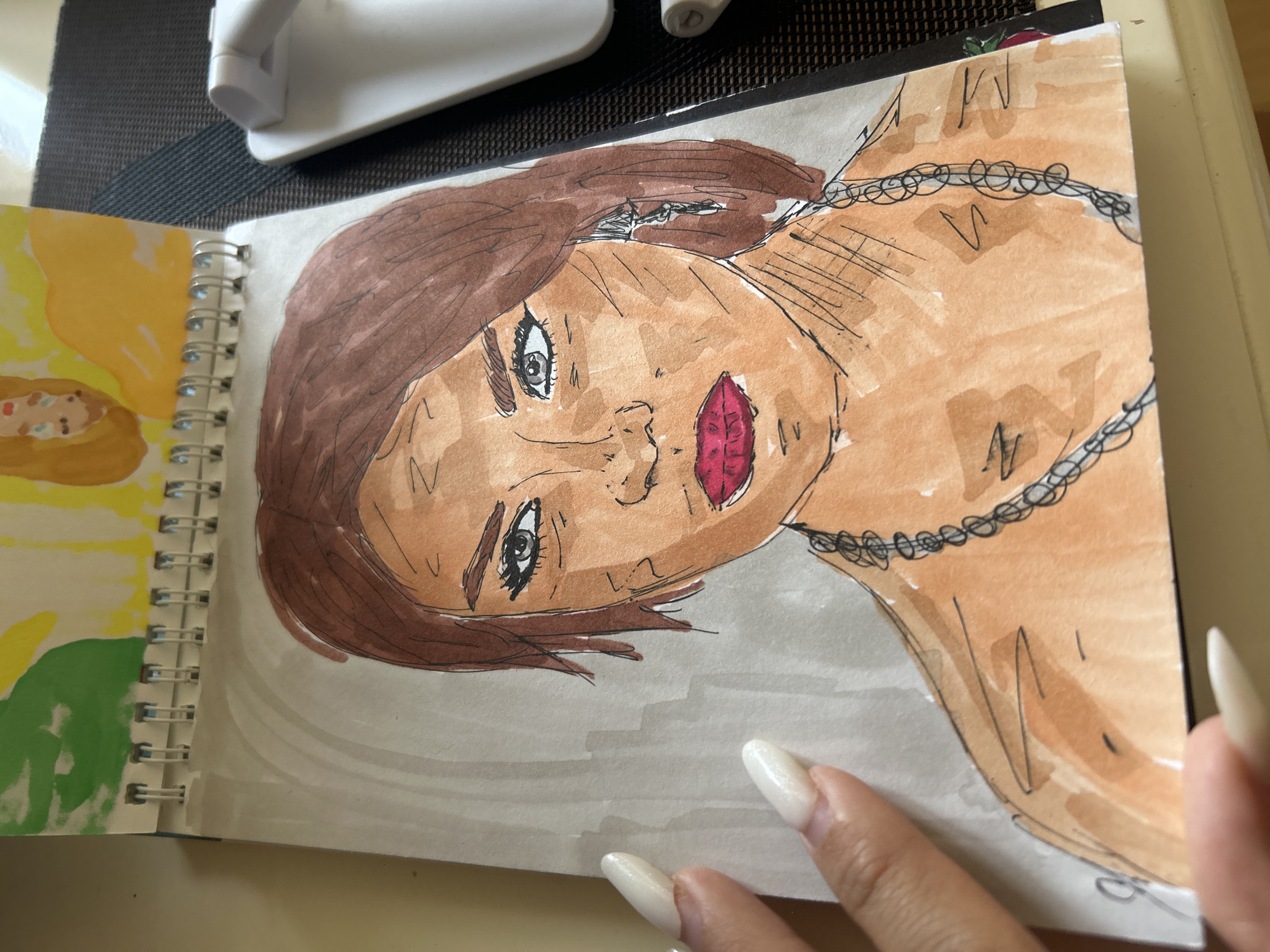 A close-up, overhead shot captures a watercolor and ink drawing of a woman's face and upper body in a spiral-bound sketchbook. The woman has brown hair, blue-grey eyes with heavy eyeliner, and bright red lips. She wears a silver necklace and an earring in her visible ear. The drawing style is stylized, with bold lines and watercolor washes for skin tone and hair. A hand with long, white fingernails is visible on the left side of the image, with fingertips touching the sketchbook. The background of the sketchbook features abstract green and yellow shapes. The sketchbook lies on a surface that appears to be a table or desk, partially covered by a dark placemat. A white electronic device with a curved handle is visible to the right of the sketchbook. The scene is well-lit, suggesting an indoor environment. The location of Khyrdalan, Azerbaijan is not depicted in the image, nor are any elements within the image that would indicate this location.