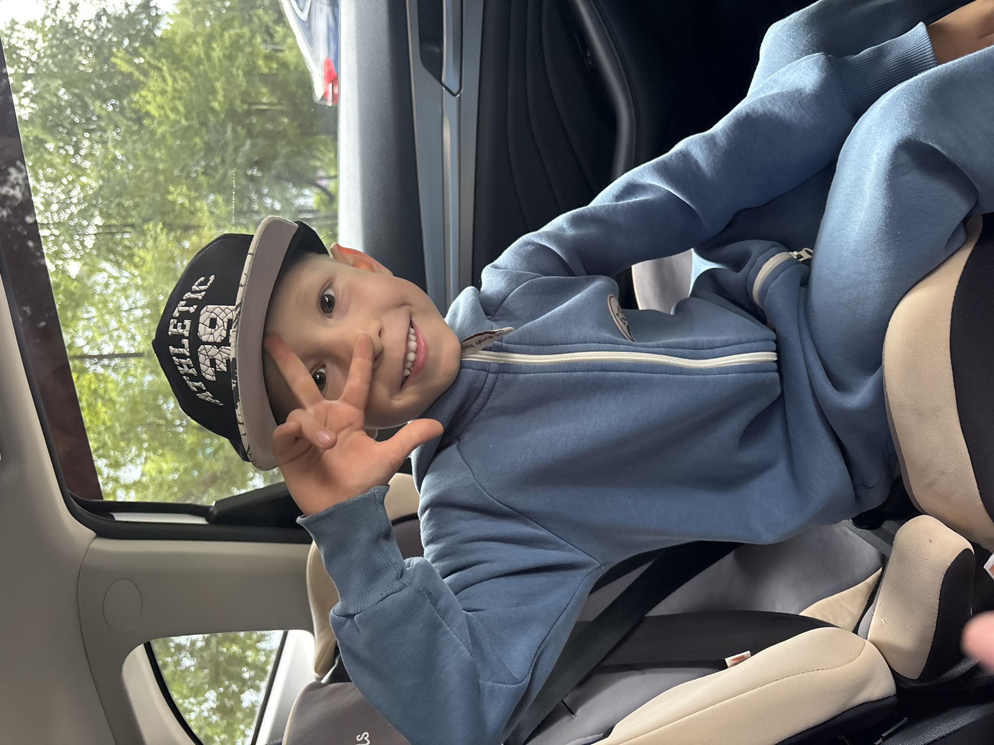 A young boy is captured in a bright, informal portrait taken inside a car, likely in Chaikovsky, Russia. He is the central subject, positioned in a car seat and angled towards the camera, smiling broadly with a playful expression. The boy is wearing a light blue zippered tracksuit and a black and grey baseball cap, tilted slightly backward. He holds up his left hand, making a peace sign gesture.
The setting is the interior of a vehicle, suggested by the car seat, visible seatbelts, and car door frame. Through the car windows, green foliage and blurred trees indicate an outdoor, daytime scene, possibly during travel. The car's interior features light-colored trim and a dark grey or black car seat, complemented by a visible sunroof in the upper left of the frame. The overall impression is one of a cheerful moment during a journey.
Notable details include the text "ATHLETIC 68" prominently displayed in white on the front of his black baseball cap. A small, partially visible label on the zipper pull of his tracksuit reads "hungry boy." The lighting suggests it is daytime with good natural light.