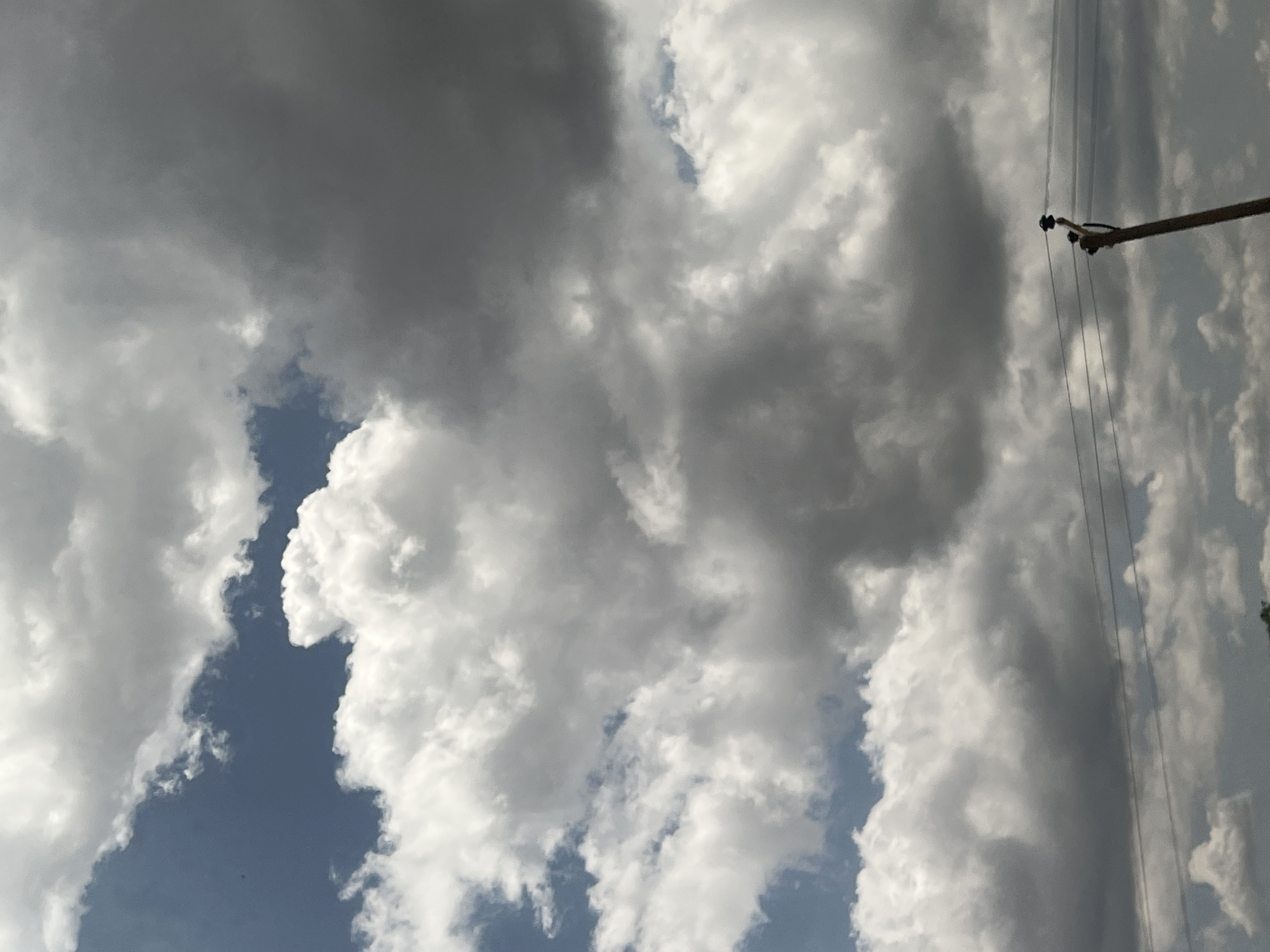 The image captures a dynamic sky scene in Sabon Layi Jessu, Nigeria, dominated by a diverse cloudscape under daylight conditions. The upper-left quadrant reveals patches of clear blue sky. The majority of the frame is filled with clouds ranging from bright, billowy white cumulus formations, particularly evident in the mid-left, to denser, darker grey clouds occupying the right side and lower-middle sections, suggesting significant atmospheric moisture or varying cloud thickness. In the lower-right foreground, the upper section of a wooden utility pole is visible, angled slightly, with a black insulator at its peak. Three thin, dark power lines extend horizontally from this pole across the lower-middle portion of the image. No human figures, animals, or specific actions are depicted. The overall environment appears to be an open sky view, with minimal ground features visible only at the very bottom edge, possibly indicating distant foliage.