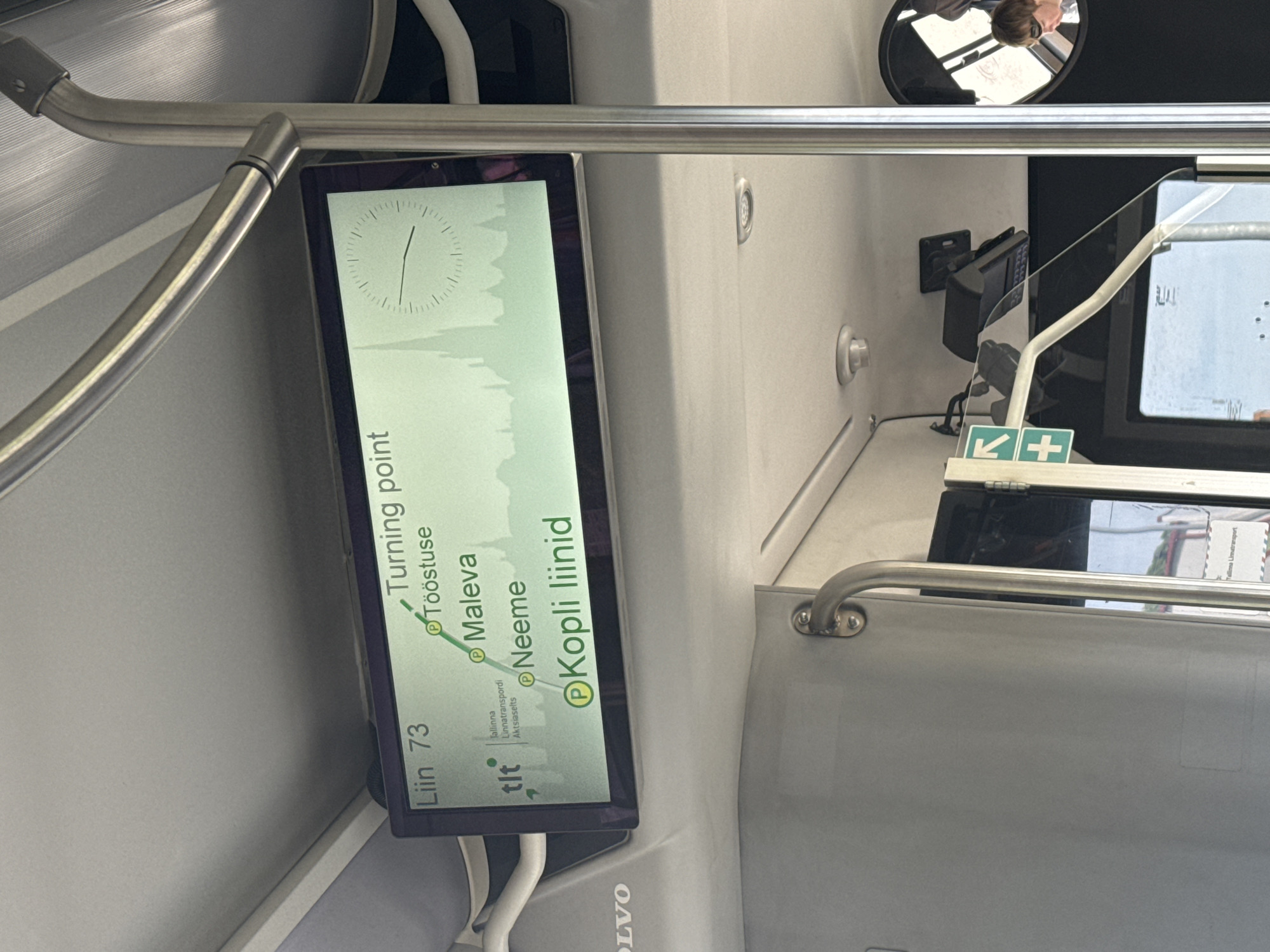 The image shows the interior of a Tallinn public transport bus, likely a trolleybus, with a digital display showing route information. The display indicates "Liin 73" (Line 73) and lists upcoming stops: "Turning point", "Tööstuse", "Maleva", "Neeme", and "Kopli liinid". A stylized silhouette of Tallinn's skyline is visible in the background of the display, along with a clock face. The "tlt" logo, representing Tallinna Linnatranspordi Aktsiaselts (Tallinn City Transport Company), is also present on the display. Metal support poles and a grey interior are visible. A sign with a green cross and arrow is mounted on a pole to the left of the display, and a small black device, possibly a ticketing machine or camera, is mounted above the display. Part of a person wearing sunglasses is reflected in a circular mirror on the right side of the image. The overall lighting suggests it is daytime.