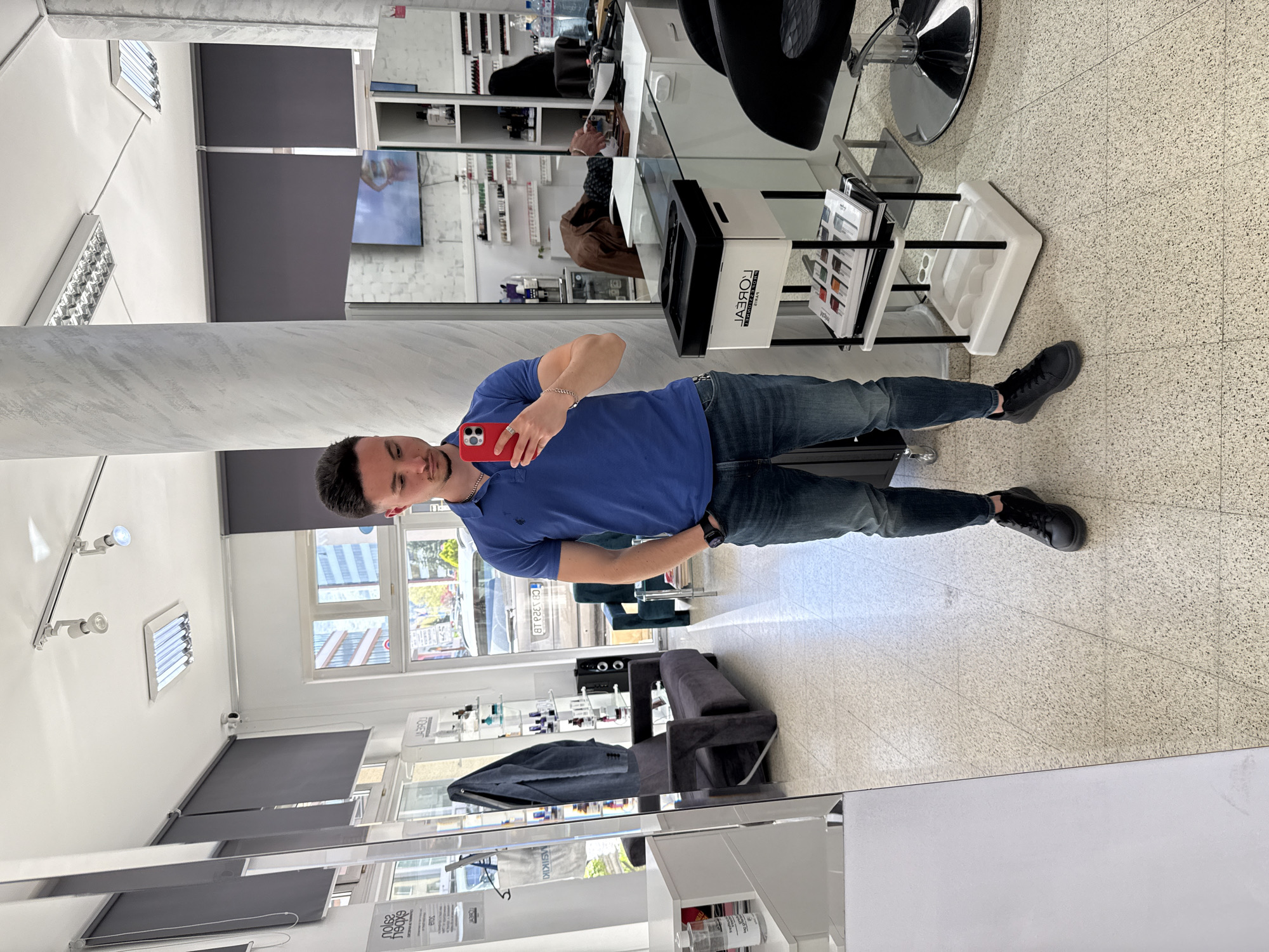 The image captures a selfie taken by a young to middle-aged man in a large salon mirror, appearing rotated 90 degrees clockwise from its natural orientation. The man, with short dark hair, is wearing a blue polo shirt, dark blue jeans, and black sneakers. He has a watch on his left wrist and a chain bracelet on his right, and holds a red smartphone (likely an iPhone) to take the picture.
The setting is a modern hair salon, characterized by a light-colored speckled terrazzo floor, white walls, and a white ceiling equipped with strip lighting and spotlights. Large windows are visible through reflections, showing an urban street scene with cars (one license plate partly visible as "CB 323..."), buildings, and trees, indicating daytime. Window blinds are partially drawn.
Reflected in the mirror are various salon elements: black adjustable salon chairs with chrome bases, white shelving units displaying numerous hair care products (bottles, tubes), a white rolling cart, and a small dark grey sofa. A white rectangular box on a shelf prominently features the brand name "L'ORÉAL" (reflected backward). In the background, a flat-screen TV is visible, displaying an image of a person. A marble-patterned column is also part of the salon's architecture. The overall impression is a clean, well-lit professional environment.