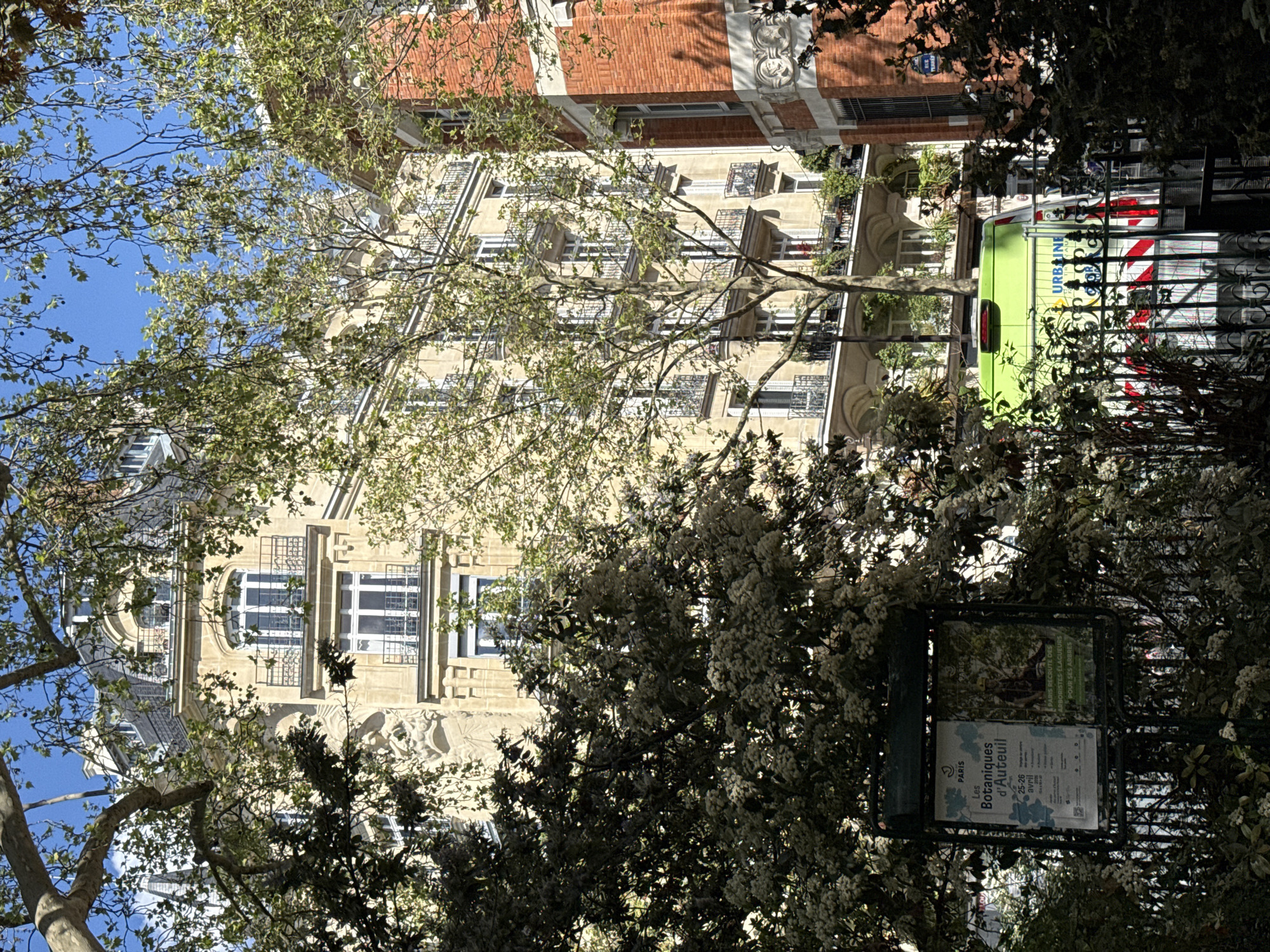 The image depicts a street scene in Paris, France. A multi-story building with ornate architectural details and numerous windows is partially visible behind trees. A bus is parked alongside the building. A metal sign is in the foreground, with the text "Les Botaniques d'Auteuil" and the dates "25-26 Avril" visible.
