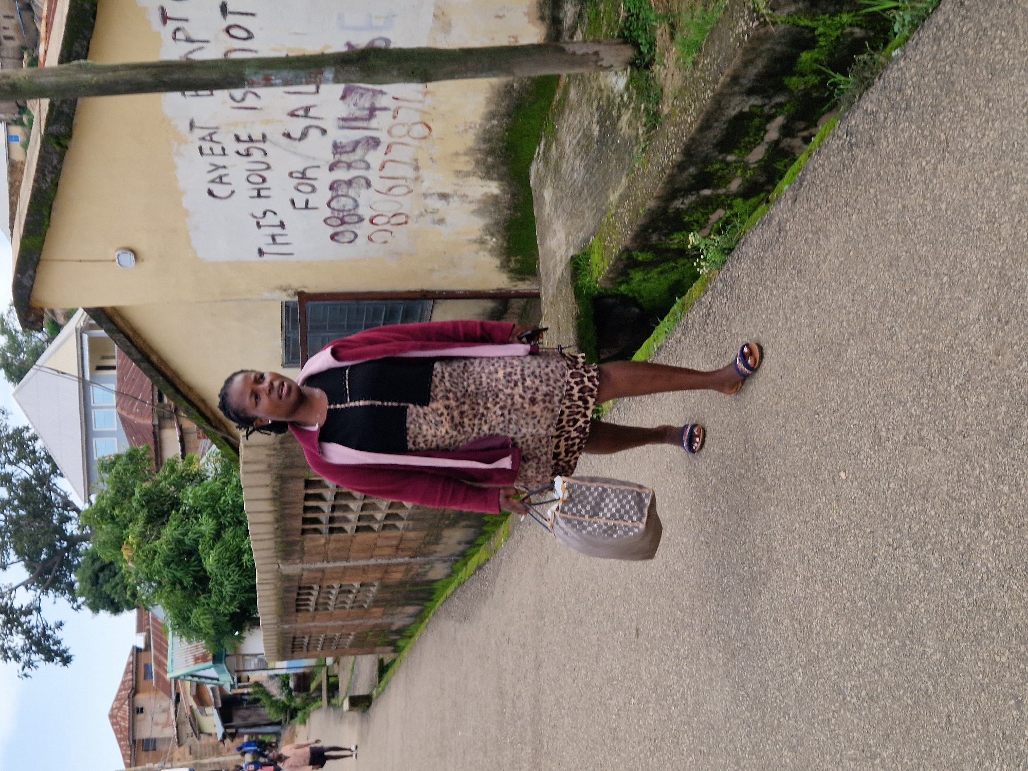 A woman walks on a paved road in Kabong, Nigeria, carrying a patterned duffel bag. She wears a maroon jacket over a black top and a leopard-print skirt, with dark sandals on her feet. The road is lined with a concrete fence on the left and a yellow building with graffiti on the right. The graffiti includes writing such as "CAY EAT BAPT" and "THIS HOUSE IS NOT FOR SALE" followed by phone numbers. The environment appears to be a residential area with some vegetation and other buildings visible in the background. A tree trunk stands near the building with graffiti. Another person is walking in the distance on the left side of the road.