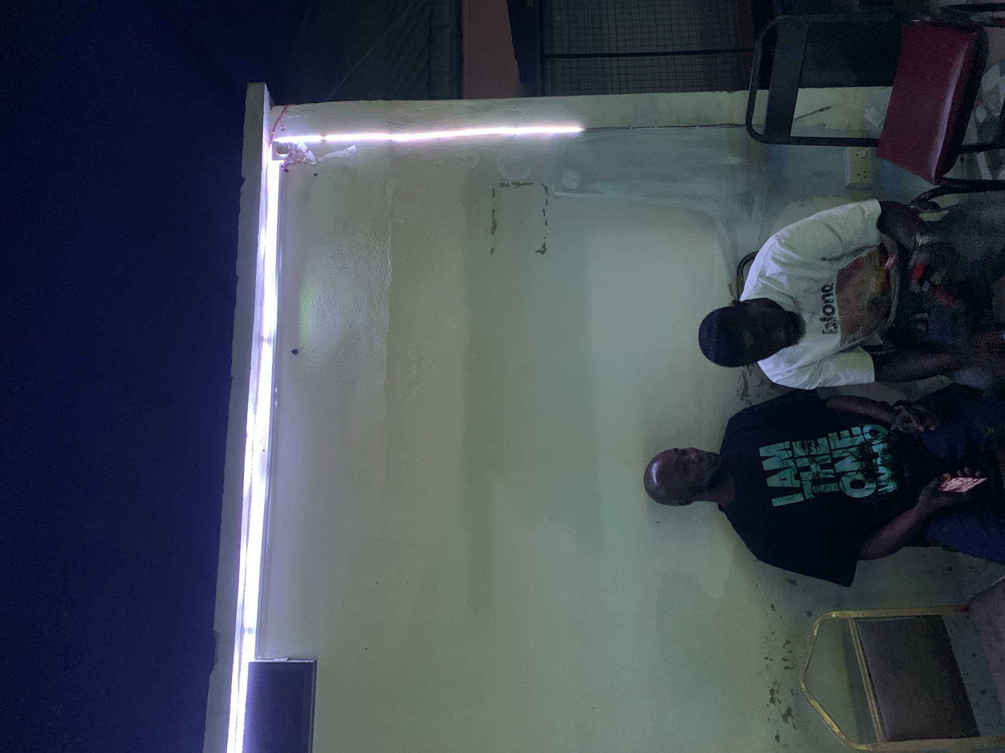 Two men are seated outside a building at night. One man wears a black T-shirt with a green graphic print and holds a phone. The other man wears a white T-shirt. They are in New Stadium, Nigeria. The building has a bright white light fixture along its top edge. A red-backed chair is visible.