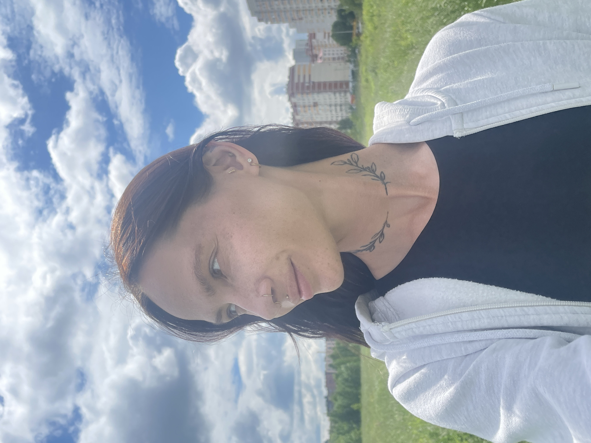 A mid-shot photograph shows a woman in profile, facing left, outdoors in a grassy field with a blue sky filled with white clouds. She has long, dark hair, fair skin, and is wearing a black shirt and a white zipped hoodie. She has several piercings: a septum piercing, a labret piercing, and earrings. A delicate branch tattoo is visible on her neck. In the background, there are trees and several multi-story residential buildings. The overall scene suggests an urban fringe environment.