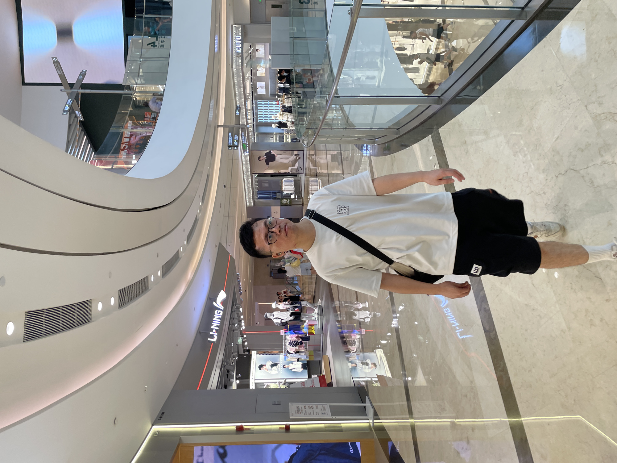 A young East Asian man wearing glasses, a white t-shirt, and black shorts is walking through a modern indoor shopping mall. He has a black crossbody bag slung over his right shoulder. The mall features a curved, multi-level architectural design with a white ceiling that has integrated lighting and air vents. The floor is a polished marble with inlaid darker lines. In the background, there are various retail storefronts, including a Li-Ning store, with mannequins displaying clothing. People can be seen shopping and walking in the distance. The scene appears to be in the Yuzhong District of China, judging by the mall's architectural style and signage.