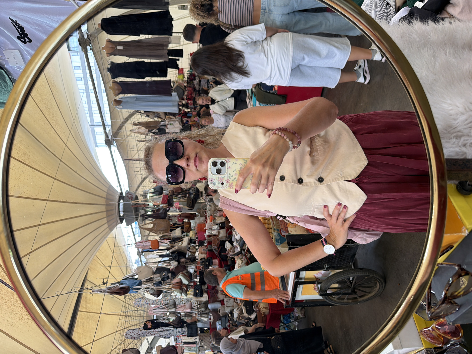 The image is a reflection in a round, gold-rimmed mirror. The mirror captures a busy outdoor market scene, likely in Kadıköy, Turkey, evidenced by the diverse crowd and variety of goods. In the foreground of the reflection, a woman is taking a selfie with her phone. She is wearing sunglasses, a light-colored sleeveless vest over a patterned shirt, and a long, deep red pleated skirt. She has several bracelets on her left wrist and a watch. Her phone has a floral case with a light yellow background.
Behind the woman, the market stalls are visible. There are many handbags and clothes hanging from racks. In the middle ground, a man wearing an orange safety vest and a blue shirt is seen interacting with a display of bags. To his left, a woman in a grey headscarf and black dress is also looking at the goods. Further in the background, other shoppers are browsing the stalls. The market appears to be covered by a large, light-colored canopy. The overall atmosphere is that of a bustling marketplace with a variety of retail offerings.