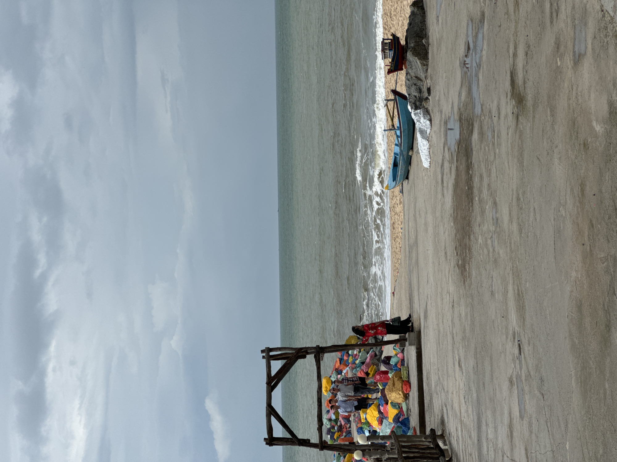 This is a photograph taken at a beach in Phan Thiết, Vietnam. The scene depicts the ocean and beach under a cloudy sky. In the foreground, a concrete walkway extends towards the beach. To the left, there is a wooden structure with colorful rocks piled beneath it. Several people are standing near the rocks and on the walkway. Further to the right, two boats are resting on the sandy beach near the water's edge. The water is choppy, with waves breaking on the shore. The overall atmosphere is overcast.