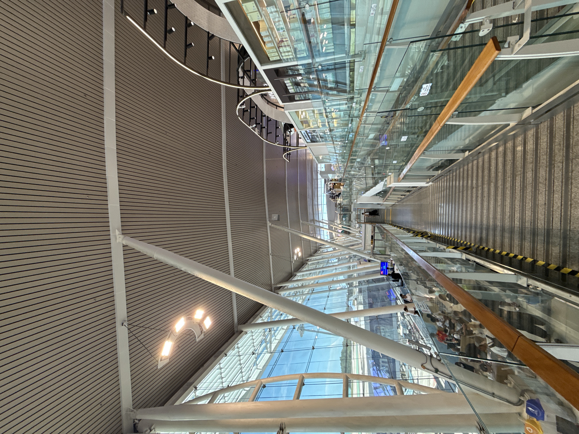 The image shows an interior view of a modern airport terminal, likely in Tambon Mai Khao, Thailand, given the context. The perspective is from a walkway or elevated corridor, looking down a sloped path with a moving walkway or escalator. Large glass windows line the left side of the corridor, offering a view of an airport tarmac with planes visible in the distance, as well as a clear blue sky with some clouds. The architecture features a high, slatted ceiling with prominent white support beams and integrated lighting fixtures.
On the right side of the corridor, a multi-level structure is visible, featuring shops or retail spaces with glass fronts displaying merchandise. This structure is also lined with glass balustrades and illuminated with curved LED lights.
In the foreground and midground, numerous people are present, walking along the corridor, waiting, or seated in designated areas. They are diverse in age and are dressed in casual travel attire. Some individuals are interacting with each other, while others appear to be focused on their phones or looking towards the shops.
The overall environment is bright, clean, and organized, typical of a contemporary airport. The materials used are predominantly glass, metal, and wood accents, contributing to a sleek and airy aesthetic. The moving walkway or escalator in the foreground has yellow and black striped safety markings. Various signs and informational displays are also present within the terminal.