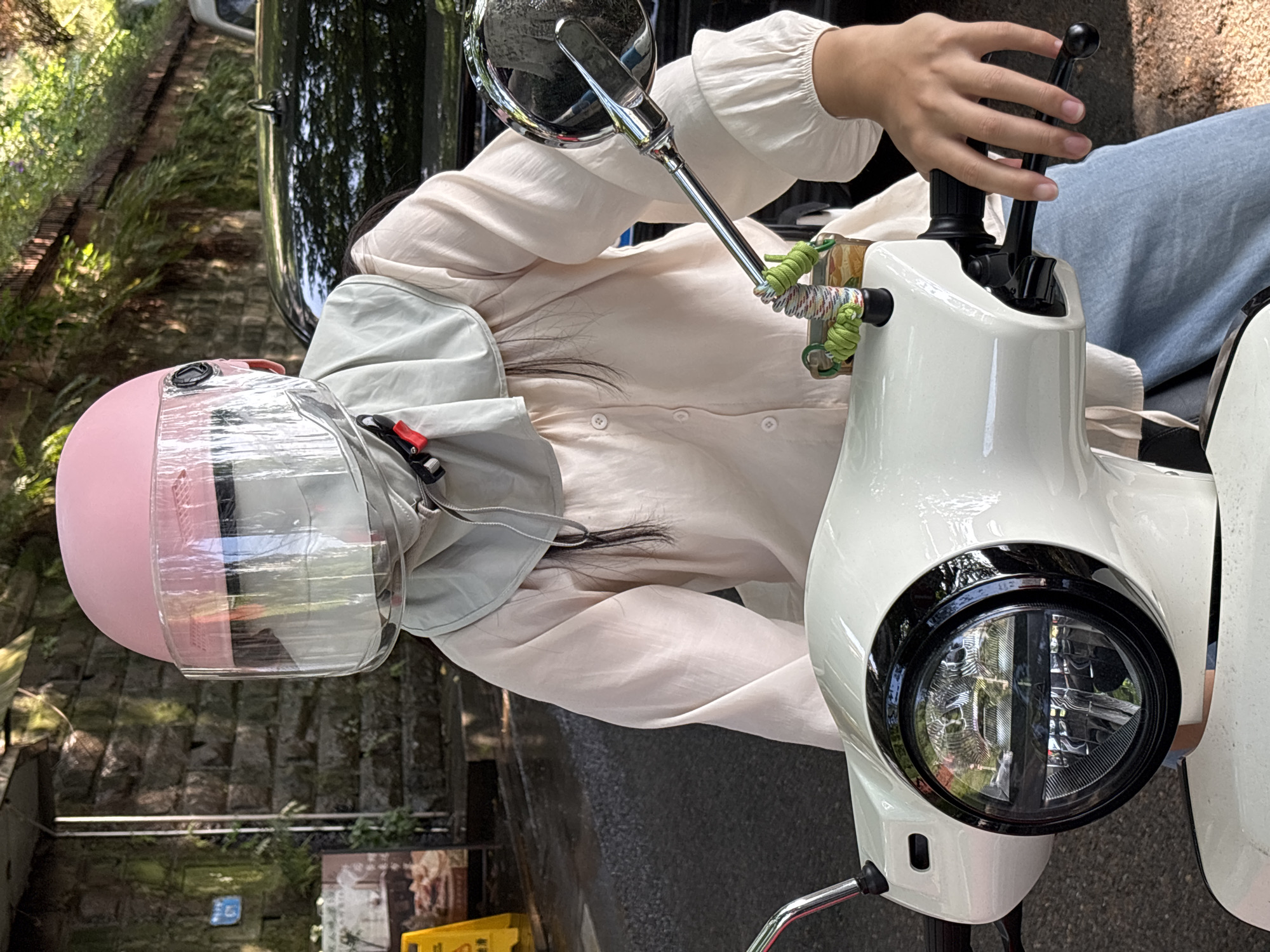 A person wearing a pink helmet with a clear face shield, a light-colored collared shirt, and jeans sits on a white moped with a black-rimmed headlight. The moped has a black handle in front of the person. A side mirror is visible on the left side of the frame. The background features a stone wall and greenery, suggesting a roadside location in a city. The surroundings include glimpses of a black vehicle and street furniture, hinting at an urban environment possibly in Yuzhong District, China.