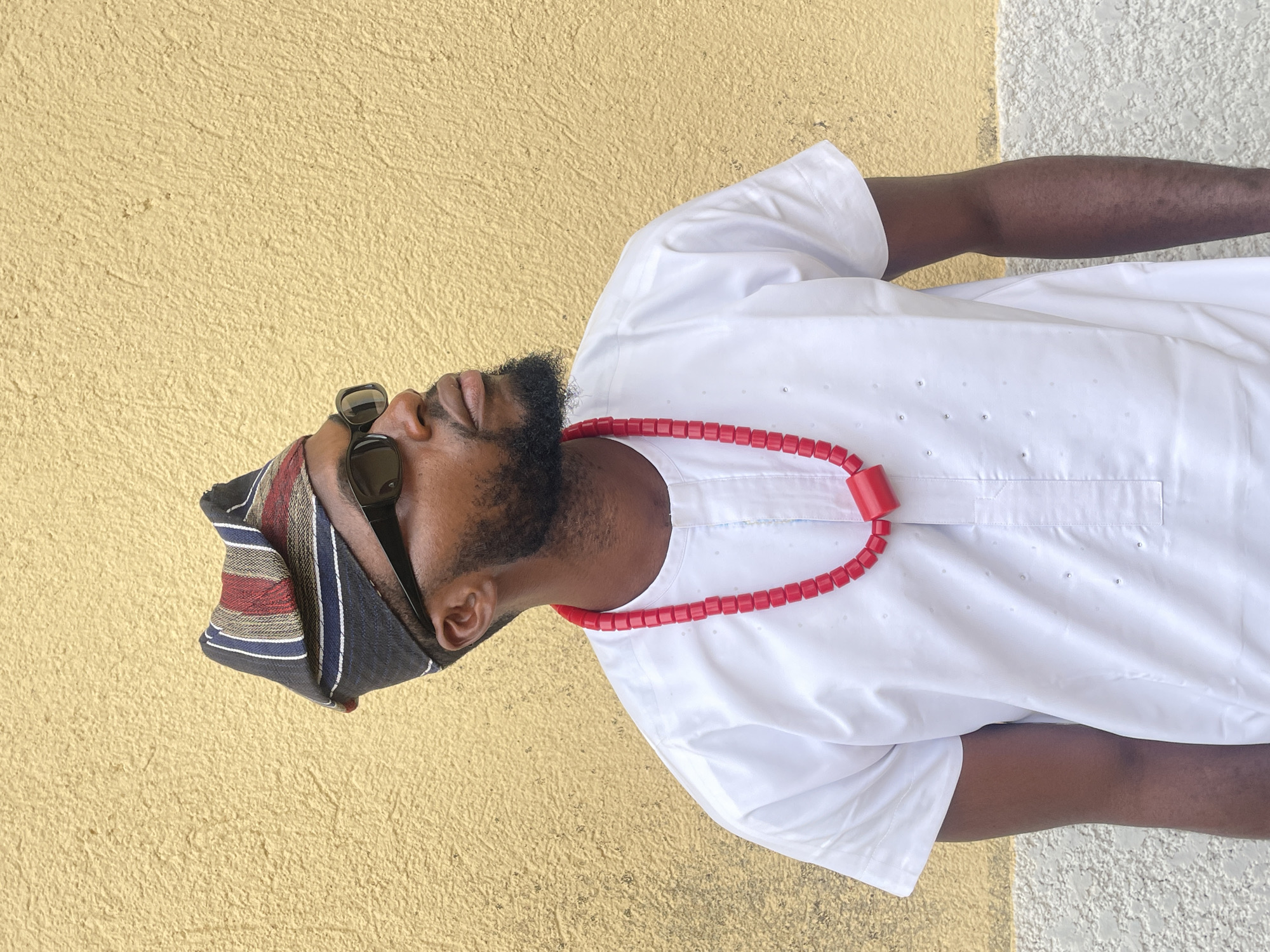 A Black man is depicted in a medium-shot portrait against a textured wall in Jos, Nigeria. He is positioned slightly off-center to the right, facing and looking upwards and to his right. The man wears a multi-colored striped cap (fila) featuring black, white, red, blue, and gold stripes. He has a full dark beard and wears black rectangular sunglasses. A long, vibrant red beaded necklace, consisting of numerous small square beads and a larger central rectangular bead, hangs around his neck. He is dressed in a plain white, short-sleeved garment, likely a traditional tunic, with a round neckline and a short placket. The background consists of a rough, textured wall; the upper portion is painted a warm, light yellow, while a lower section on the left reveals a lighter, unpainted or white-painted textured surface.