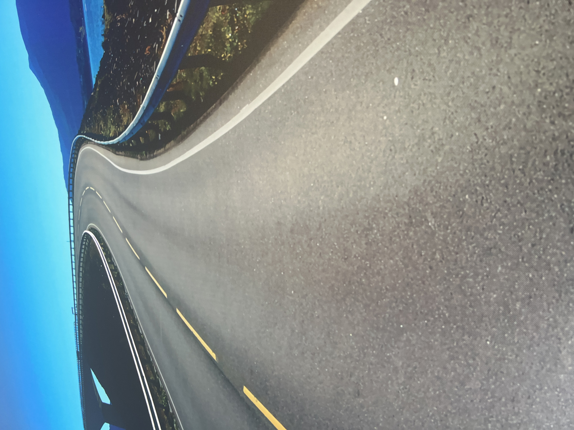 This is a photograph of a winding road with guardrails, bordered by trees and a body of water. The road has white and yellow lane markings. The sky is a clear, bright blue. The overall scene appears to be a scenic mountain or coastal road. The image is taken from a high angle, looking down at the road as it curves away. There are no people visible in the image. The lighting suggests it is daytime, likely with fair weather.