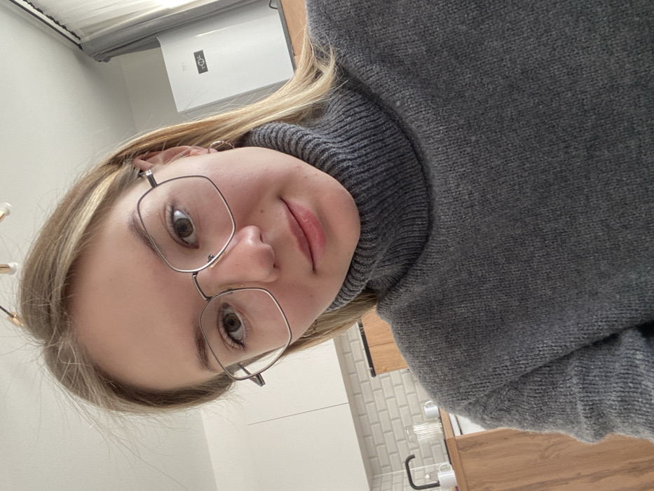 An image taken in a residential interior, specifically a kitchen area, in the city of Bittsa, Russia, depicts a single female subject in a close-up, likely a selfie. The subject, positioned centrally, has fair skin, brown eyes, and blonde hair, which is parted and extends over her shoulders. She wears large, silver-framed, hexagonal eyeglasses and a dark grey knit turtleneck sweater. A small silver hoop earring is visible in her right ear. The subject is looking directly towards the camera with a neutral to slight smiling expression.
The background shows a contemporary kitchen design. To the subject's left, white upper cabinets, a white subway tile backsplash, and light brown wooden lower cabinets or countertop are visible. A black gooseneck faucet, white mugs, and a glass container are present on the counter. To the subject's right, a white appliance, possibly a refrigerator or air conditioning unit, with a small digital display is seen. Grey curtains or drapes are partially visible next to this appliance. Overhead, a modern light fixture with multiple light sources on gold-colored arms is partially visible on a white ceiling.