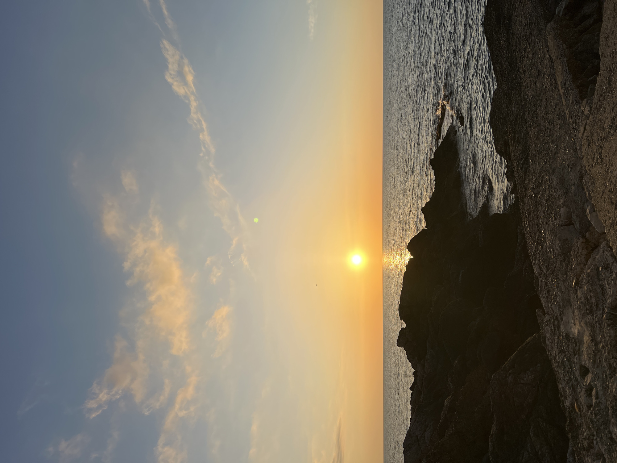 The image shows a sunset over the sea, with rocks in the foreground. The sun is a bright orange orb low on the horizon, casting a golden glow on the water. The sky is a gradient of orange, yellow, and blue, with a few wispy clouds. The sea is calm, with gentle waves lapping against the shore. The rocks are dark and jagged, suggesting a rocky coastline. There are no people or animals visible in the image.
The scene is consistent with a view from a coastal area in or near Palermo, Italy, where sunsets over the Mediterranean Sea are common. The presence of rocky shores is also characteristic of many areas along the Sicilian coast.
