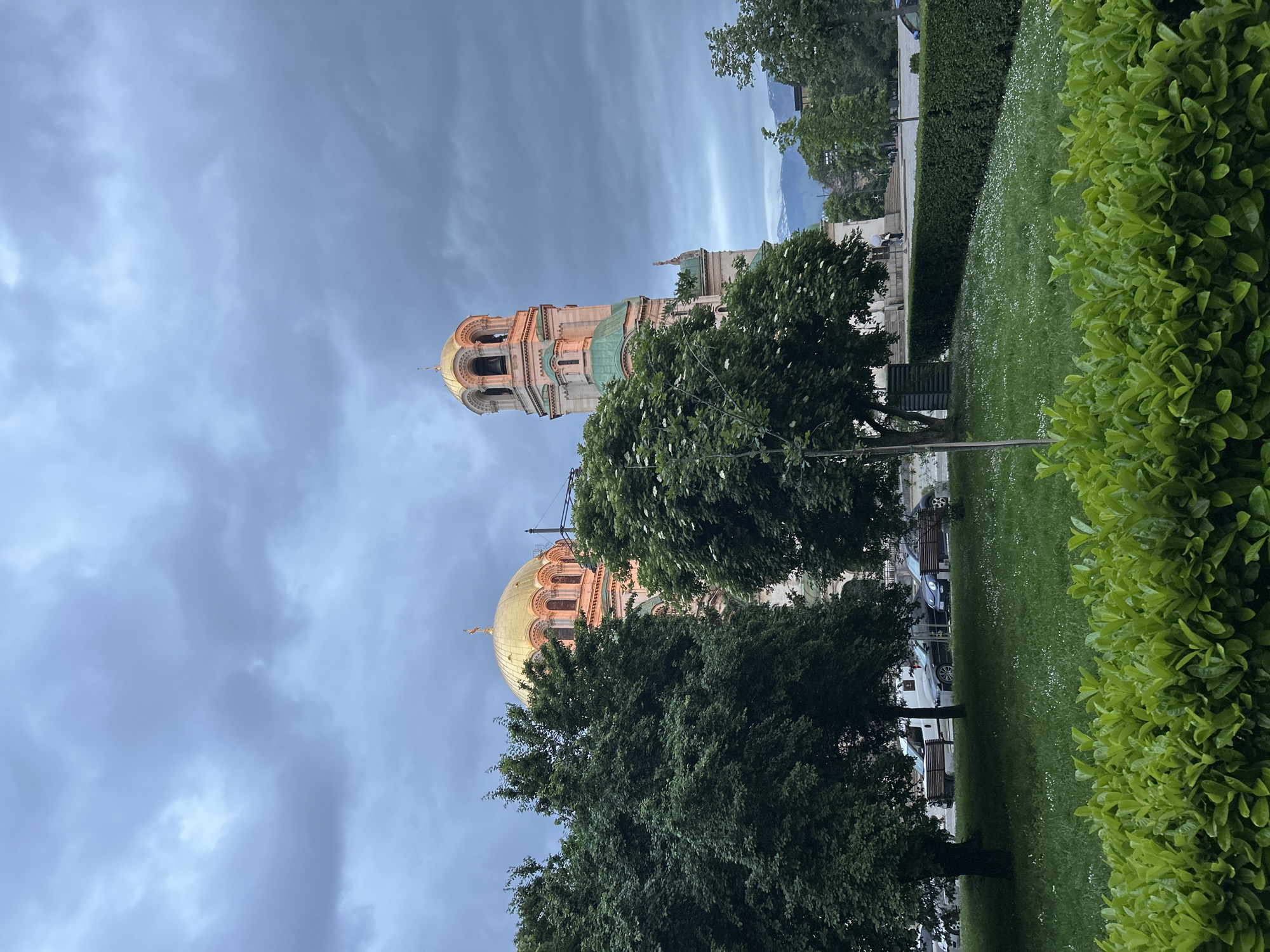 The image depicts the Alexander Nevsky Cathedral in Sofia, Bulgaria, viewed from a low angle. The cathedral's golden dome and bell tower are partially visible amidst trees and greenery. The sky is overcast. In the foreground is a green lawn with bushes on the right side. Benches and possibly cars are visible between the trees. The overall setting is urban, with elements of both natural and architectural landscapes.