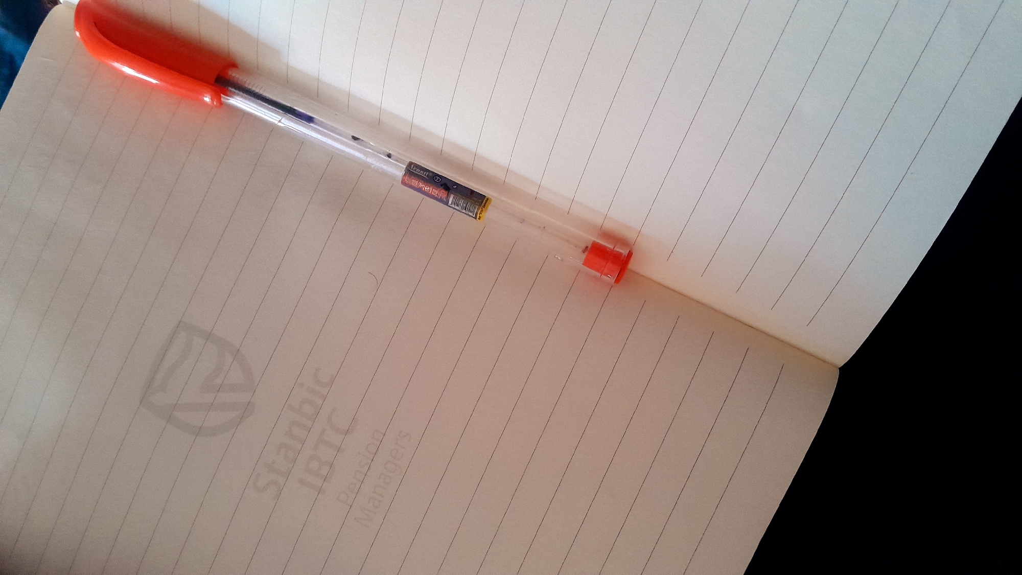 An orange and clear ballpoint pen rests diagonally across an open, ruled notebook, captured from a high-angle perspective. The pen's transparent barrel reveals a blue ink cartridge and a small white label with a barcode. Visible text on the pen's label includes "iTcraft" and "LEADER".
The notebook pages are lined with horizontal rules. The right page is largely blank, while the left page displays a faint, light grey watermark and text oriented vertically. The visible text on the notebook reads: "Stanbic", "IBTC", "Pension", and "Managers". The background around the notebook is dark, suggesting a shadowed surface. The scene is well-lit, likely indoors, with soft lighting. No active writing is occurring, but the arrangement suggests a workspace or a momentary pause from note-taking. The location context is Umuigbekwo, Nigeria.